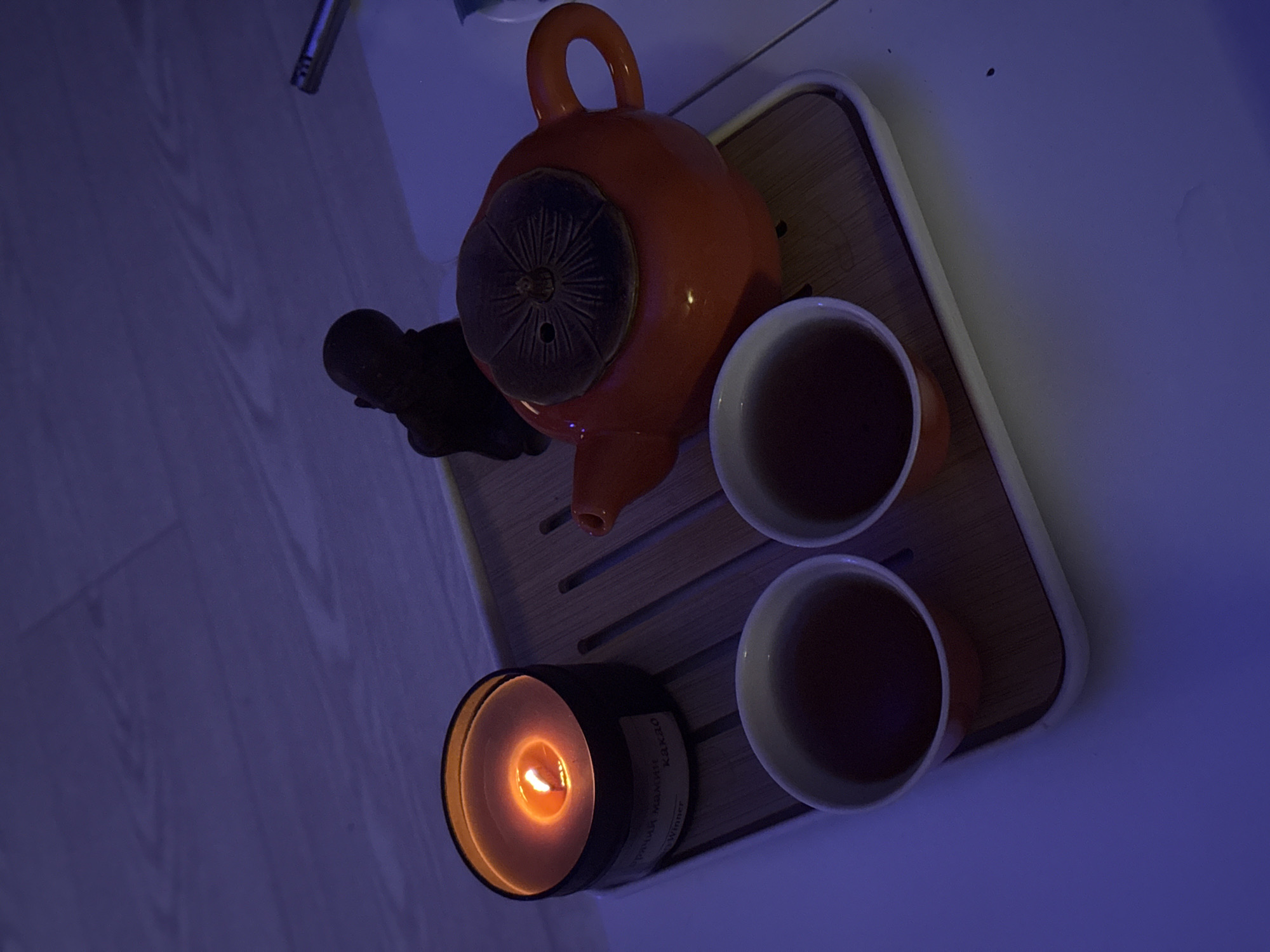 A top-down, rotated view shows an orange teapot and two orange cups filled with dark liquid on a wooden tray with white edging, placed on a light-colored surface. A lit candle in a black container is positioned near the cups. A small black statuette is next to the teapot. The label on the candle is partially visible with Russian text including "KAKAO". The image is taken indoors with mixed blue and yellow lighting. There is no evidence to place the image in Ново-Никольское, Russia.