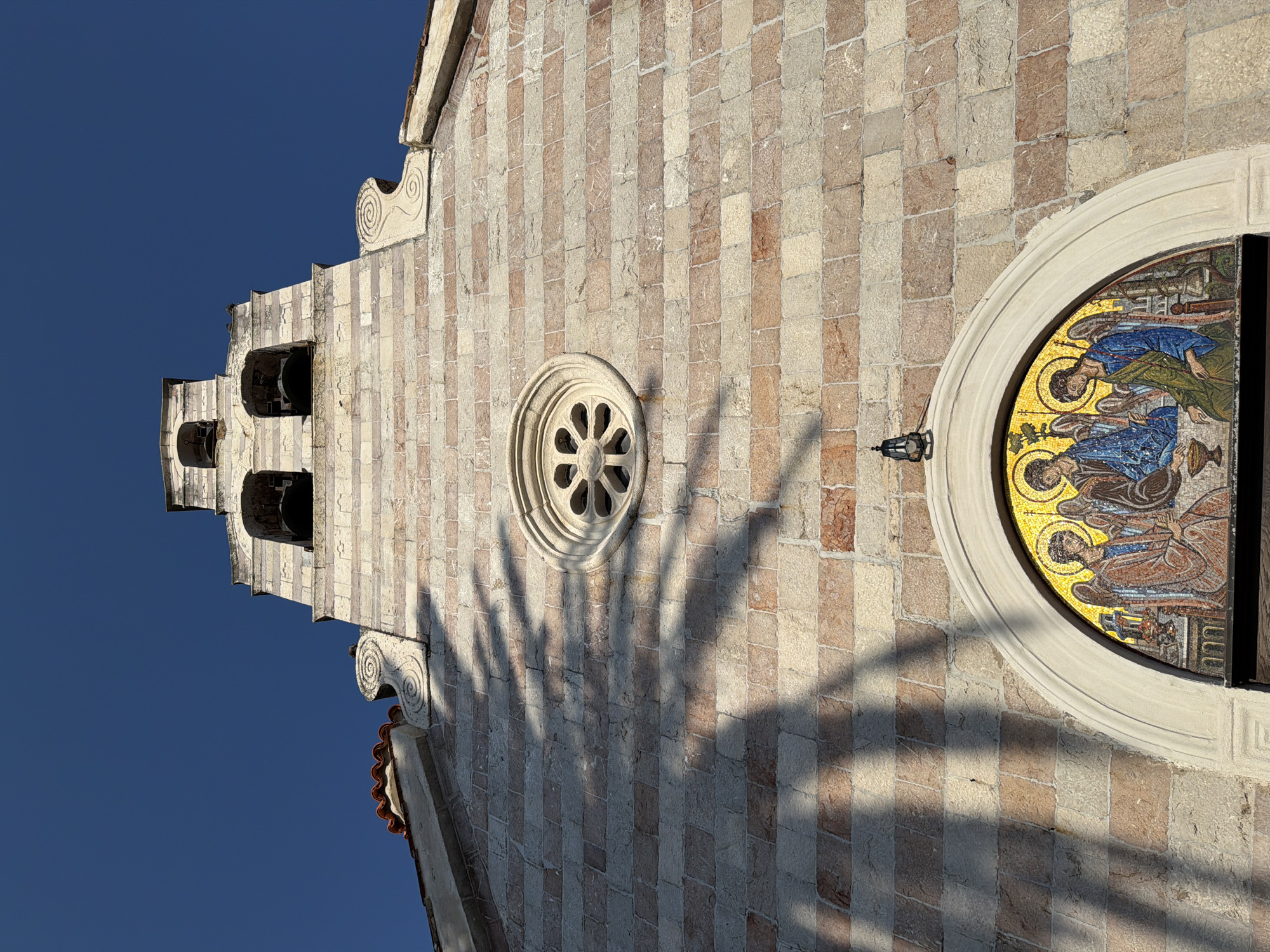 This image captures a low-angle view of the bell tower and facade of the Church of Saint John in Budva, Montenegro. The clear, deep blue sky serves as a backdrop for the stone structure, which is made of alternating bands of light and reddish-brown stone blocks. The bell tower rises towards the sky, featuring multiple arched openings with dark bells hanging within, and topped by a smaller belfry. A decorative circular window, resembling a rose window with flower-petal-like divisions, is visible on the facade. Below this, an arched mosaic depicts three figures, likely religious icons, with halos and wings, rendered in vibrant colors. The mosaic is framed by a light-colored stone arch. Shadows of what appear to be tree branches are cast diagonally across the stone facade, adding a textural element. A black lantern hangs above the mosaic, attached to the stone wall. The roofline of the church, with its red tile roofing, is visible at the top edges of the frame.
