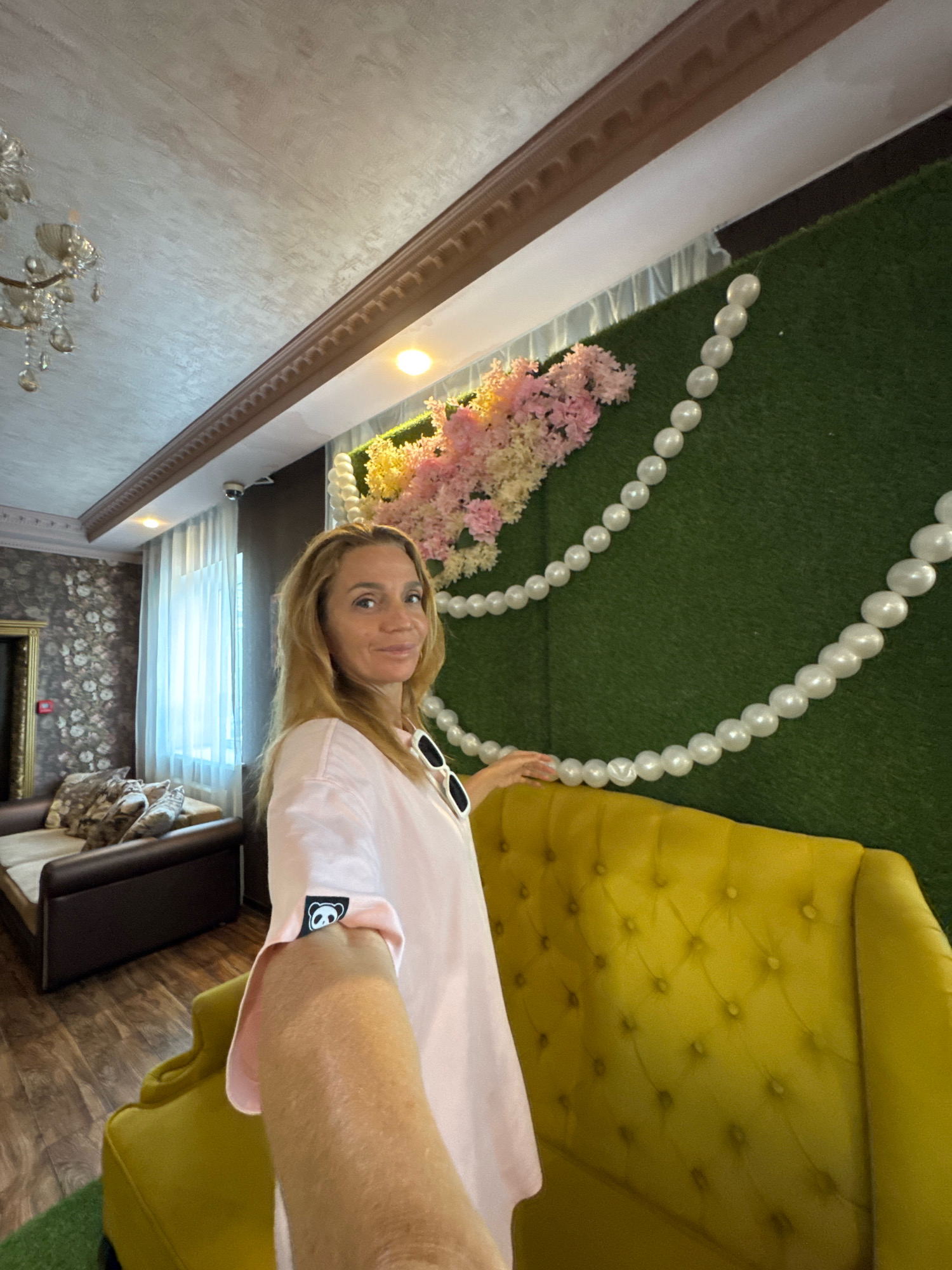 A woman with blonde hair is taking a selfie in a room. She is wearing a light pink t-shirt with a black panda logo on the sleeve. She is smiling and looking at the camera. Behind her is a green wall decorated with a string of white pearls and pink and yellow flowers. To the left of the green wall is a yellow sofa with tufted upholstery. In the background, there is a brown sofa with patterned pillows, a window with white curtains, and a chandelier hanging from the ceiling. The floor is wooden. The scene appears to be an indoor setting, possibly a salon or a decorated event space.