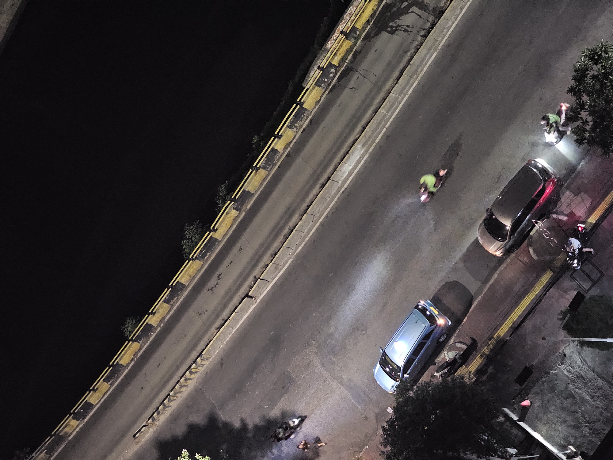 An aerial, night-time view captures a segment of a road in West Jakarta, Indonesia, likely a bridge or elevated road due to the dark expanse of water visible to the right and the road's railing. The road is bordered on the right by a yellow and black striped barrier, with concrete elements visible below. The road surface is asphalt, showing signs of wear and some dark wet patches.
Several vehicles and individuals are present. On the left side of the road, a light blue minivan is parked. Further up the road, a motorcycle with a rider is seen, emitting smoke. Near the bottom right, a dark-colored car with illuminated taillights is stopped, and two individuals on a motorcycle are nearby, with the rider illuminated by a bright headlight. In the lower left corner, a red traffic cone is visible on a sidewalk, adjacent to a tree and some structures.
The scene suggests an urban environment at night, possibly a relatively busy street given the presence of multiple vehicles and people, though the overall lighting is dim, relying on vehicle headlights and streetlights not fully captured.