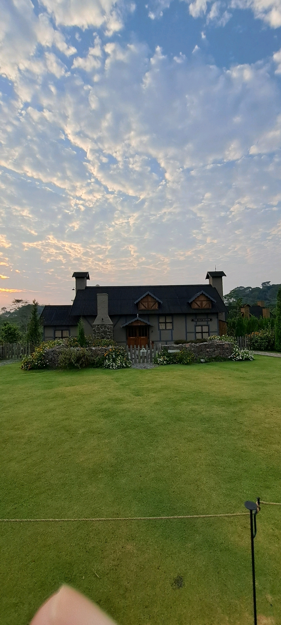 This is a vertical, eye-level, medium-shot photograph of a stone and wood manor-style house with a dark roof, situated in a grassy field. The house features two prominent stone chimneys and decorative wooden gables. A wooden fence and a low stone wall with flowering plants surround the front of the house. The foreground is dominated by a well-maintained, bright green lawn. In the background, a treed hillside and a cloudy sky with soft lighting are visible, suggesting either sunrise or sunset. A thin rope barrier is present in the lower portion of the image, and a black pole with a string attached is visible on the right. The signage on the house reads "THE OLD MANOR." The location is identified as Tamiajeng, Indonesia.