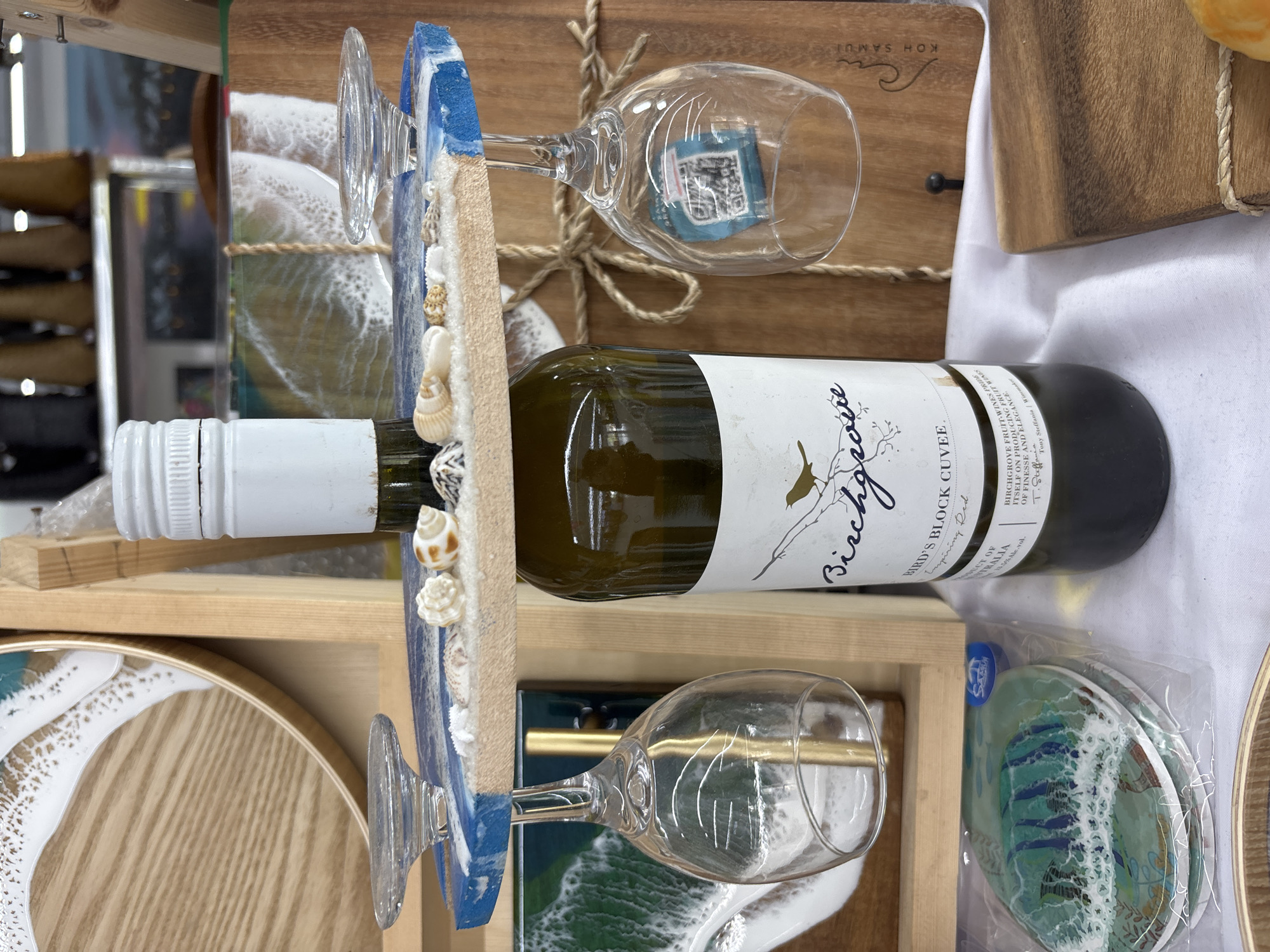 A still life featuring a bottle of "Birchgrove Bird's Block Cuvee" wine, two wine glasses, decorative beach-themed bottle/glass holder, and coasters, set against a wooden backdrop and white fabric. The scene is arranged in a wooden box. The OCR text on the bottle indicates the wine is a product of Australia, reflecting "Birchgrove Fruit-Wines," and includes the text "KOH SAMUI".