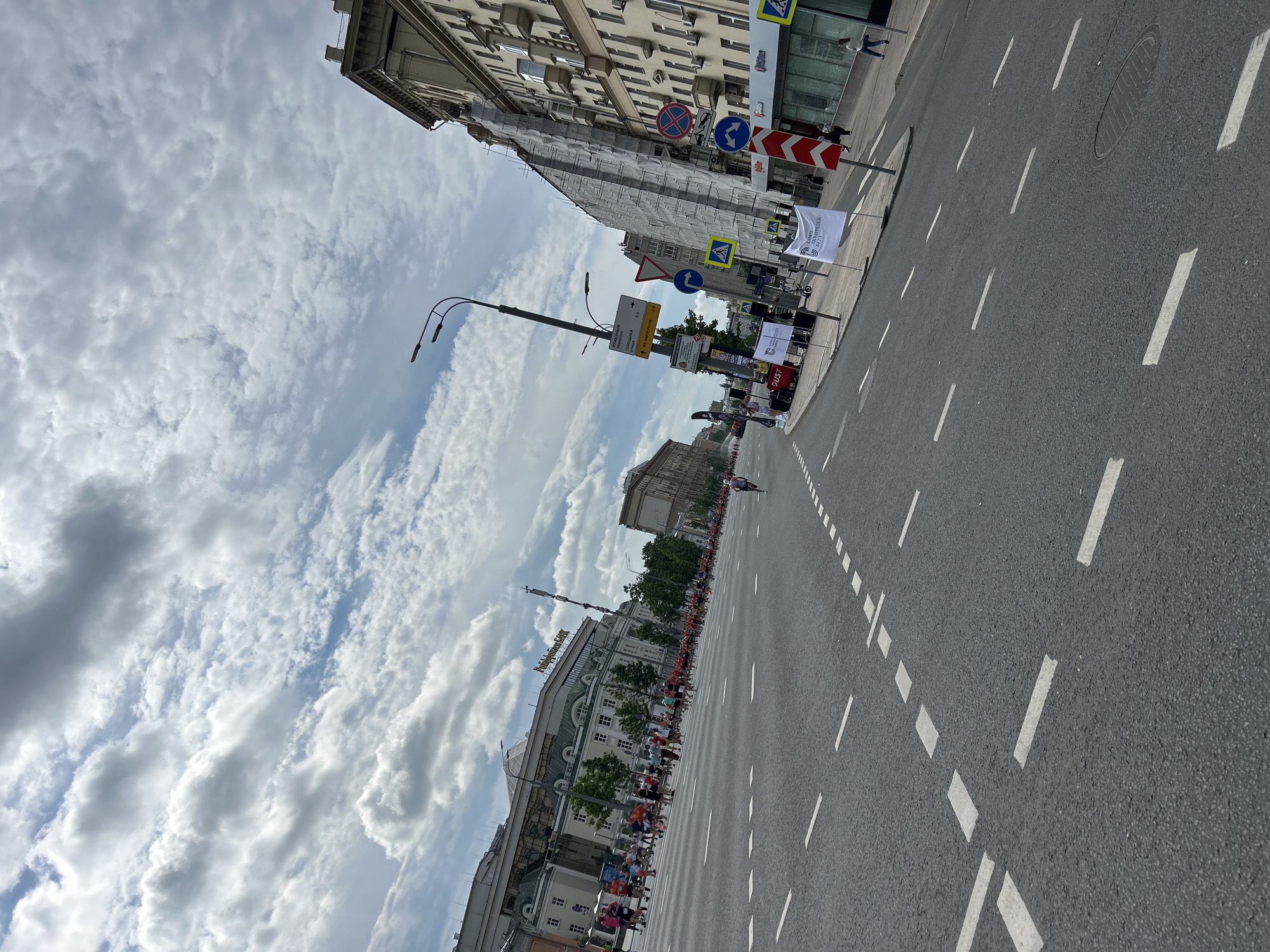 The image shows a street scene in an unknown city. The street is paved with asphalt and marked with white lines. Buildings line both sides of the street, some under construction or repair with scaffolding visible. There is a crowd of people lining the side of the road. Several road signs and banners are visible along the street. The sky is cloudy.