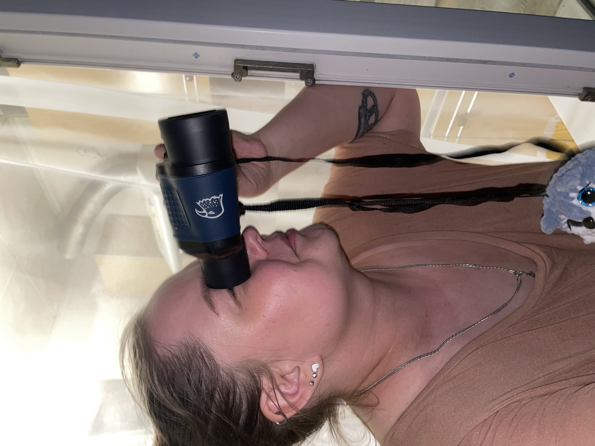 A woman is pictured from an upside-down perspective. She is holding a dark blue binocular against her forehead, positioned between her eyes. The binoculars have a strap around her neck. She is wearing a light brown shirt and a thin silver necklace. There is a blue stuffed animal visible near her shoulder. She has a tattoo on her left arm and multiple earrings in her left ear. The scene appears to be taken from within a bright, white-walled room or enclosed structure, possibly with a window or frame visible above her. There is no information to suggest a connection to the city of Izhevsk, Russia.