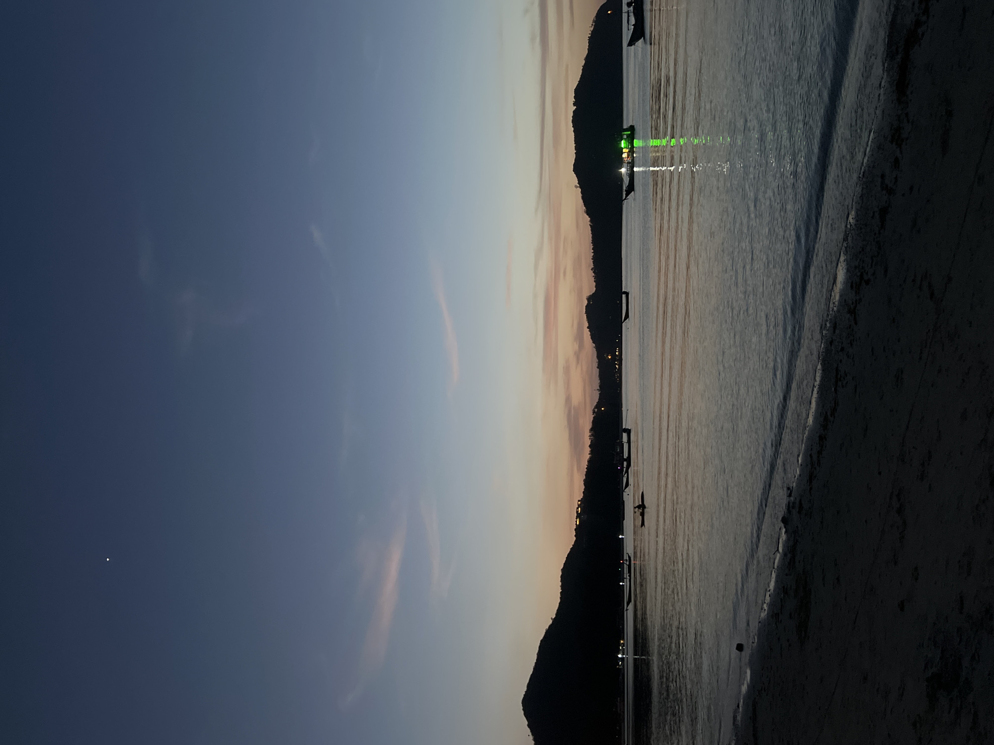 This image depicts a twilight scene on a beach in the Ko Pha-ngan District, Thailand. The sky is a gradient of deep blue at the top, transitioning to soft pinks and oranges near the horizon. A single bright star is visible in the upper left portion of the sky. Silhouetted against the horizon are the dark shapes of hills or mountains.
The calm sea reflects the muted colors of the sky and the lights from several boats anchored offshore. One boat in the mid-right section of the image is illuminated with bright green lights, casting a vibrant reflection on the water. Other boats are visible as darker shapes, some with faint lights. In the foreground, the dark sand of the beach slopes gently towards the water, with small waves lapping at the shore. The overall atmosphere is tranquil and serene.
