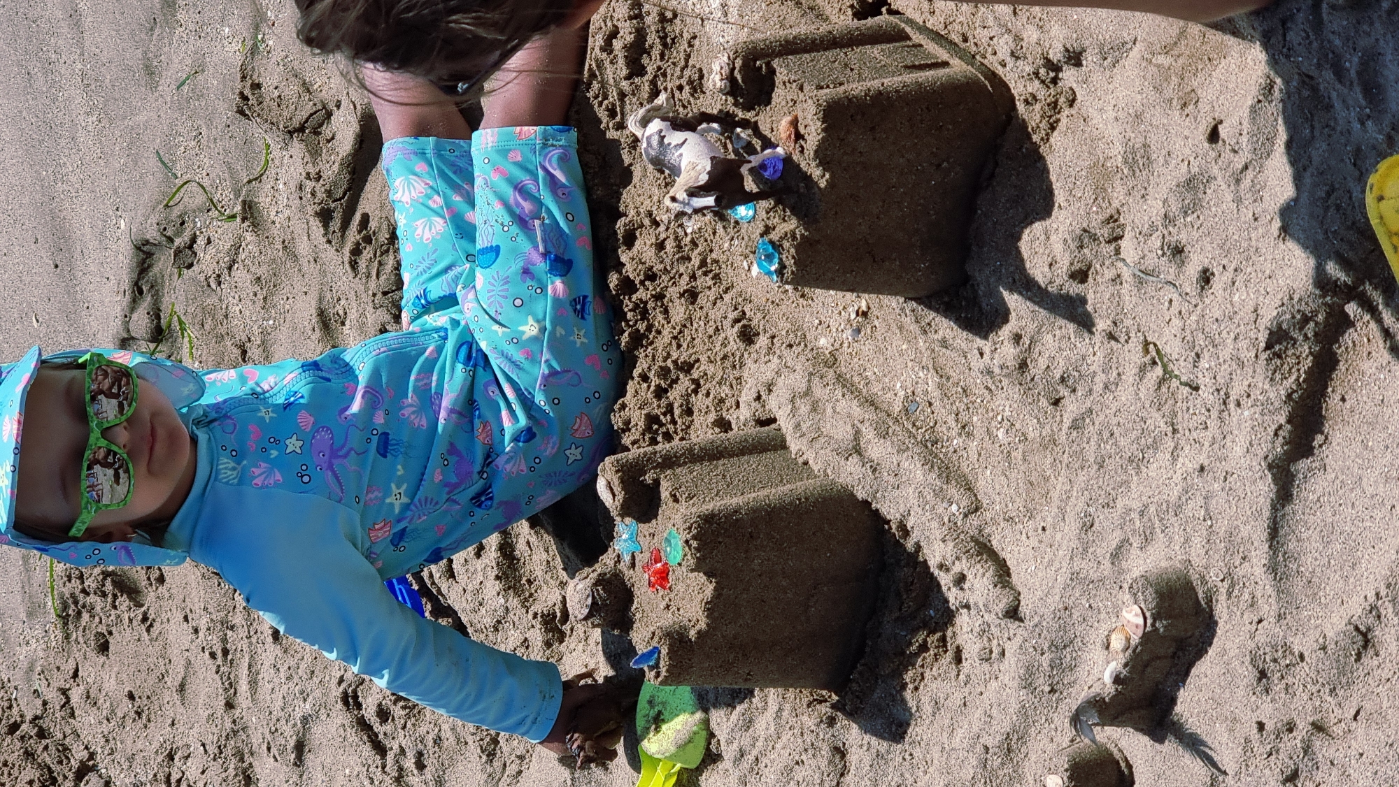The image shows a close-up, eye-level view of a young child sitting on a sandy beach, likely in Castelló d'Empúries, Spain, given the context of this query. The child is wearing a light blue rash guard with a hood, adorned with a playful sea creature print in purple and pink. Bright green sunglasses with a pattern reflect the beach scene. The child's hands are slightly blurred, suggesting they are actively playing with sand.
In front of the child are two sandcastles. The castle on the left is larger and more elaborate, topped with colorful plastic gems and what appear to be small seashells. The castle on the right is smaller and is adorned with a toy horse figurine and more plastic gems. The sand is a light brown, typical of a beach environment, with some dried seaweed scattered across the surface. The lighting suggests it is a sunny day. The lower right corner of the image shows a portion of another person's body, indicating the presence of at least one other individual. The scene depicts a typical beach activity of building sandcastles.