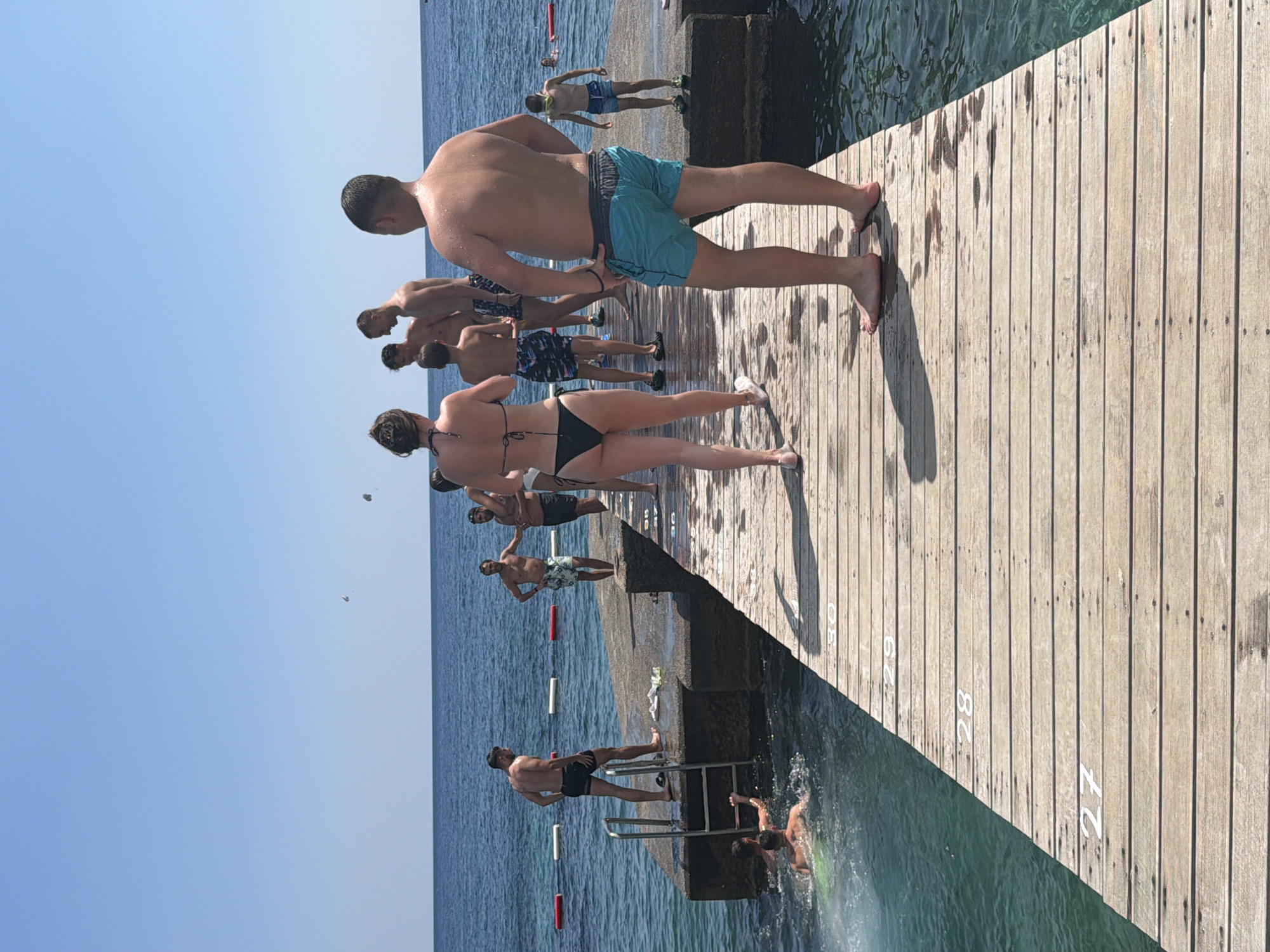 The image depicts a wooden pier extending into clear blue water, likely in Budva, Montenegro. Several individuals are visible on and around the pier. On the pier, a group of people in swimwear are standing, some facing the water and others looking away. One person is wearing a black bikini, another is in blue swim trunks, and several others are in swim shorts of various colors. Two individuals are seen in the water near the base of the pier, with one appearing to swim or prepare to jump. Another person is climbing a ladder attached to a concrete structure at the edge of the water. In the background, the sea stretches to the horizon under a clear blue sky, with two parasails visible in the distance. The pier itself has numbered markers on its wooden planks, with "27", "28", "29", and "30" clearly visible. The environment suggests a sunny day at a popular swimming or diving spot.
