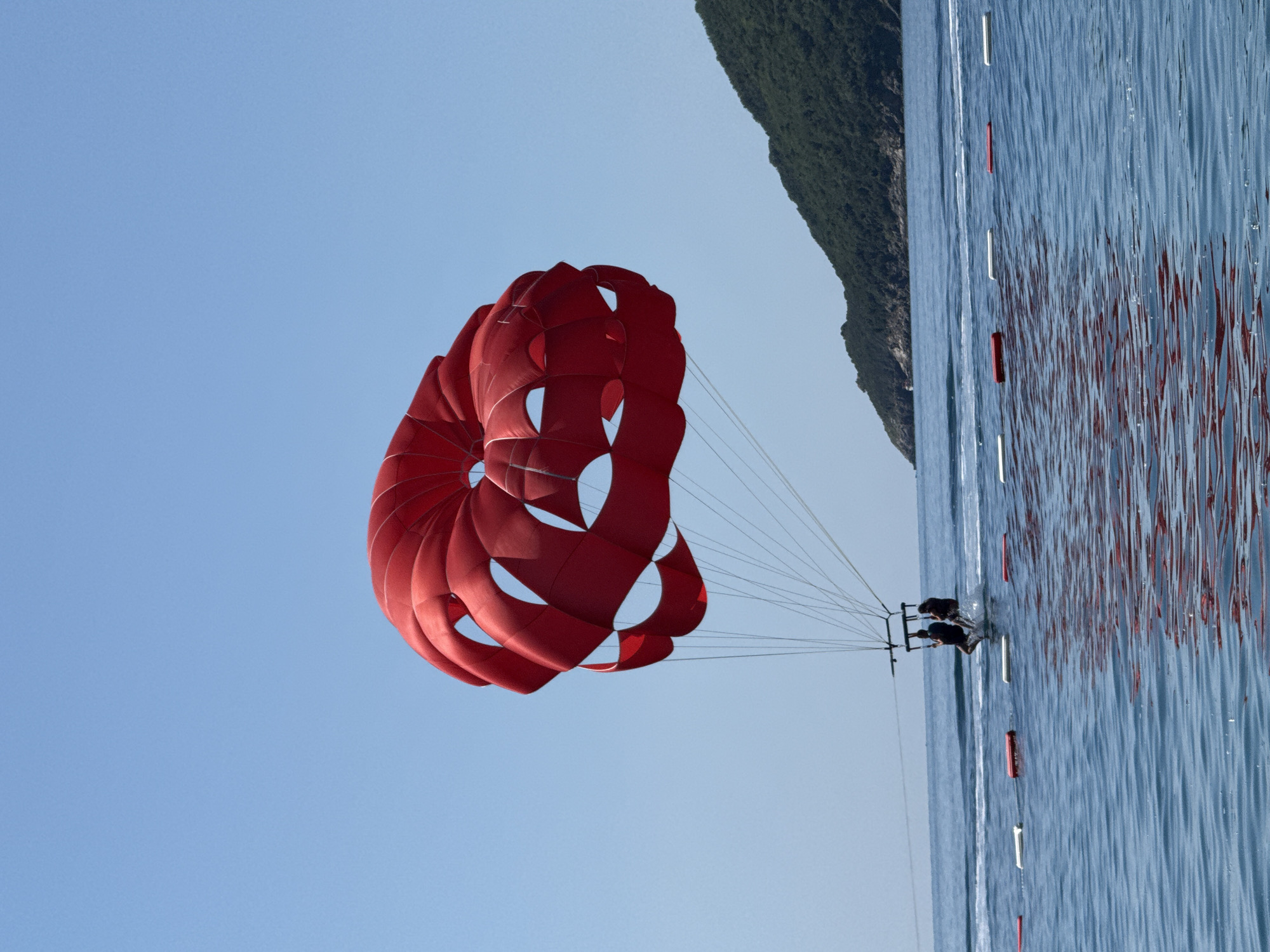 A wide shot captures two individuals parasailing over the Adriatic Sea off the coast of Boreti, Montenegro, under a clear, bright blue sky. A large, vibrant red parasail dominates the upper center of the frame, its multi-chambered canopy exhibiting varying shades of red due to light and shadow. Numerous thin white suspension lines extend downwards from the parasail to a horizontal bar, from which two people are suspended in harnesses. These individuals, seen from behind, are positioned just above the water's surface, creating a distinct splash.
The ocean water in the foreground and mid-ground is a deep blue, exhibiting ripples and reflections, notably a reddish hue reflecting from the parasail. A white, foamy wake from an unseen towing vessel extends horizontally behind the parasailers. A sequence of six oblong buoys, alternately red and white, floats on the water, forming a line across the lower mid-section of the image, demarcating an area. In the background, a verdant, tree-covered mountain or large hill defines the coastline, sloping from the right side towards the distant horizon, which features a clear blue sky meeting the water. The overall impression is one of a bright, sunny day with active watersports.