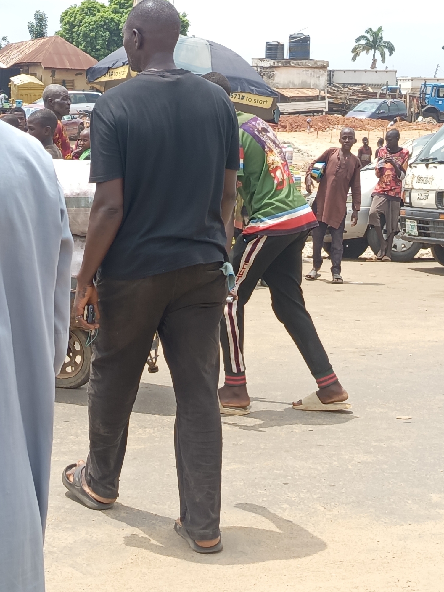 The image captures a street scene in Akwanga, Nigeria. A group of people are present. The main subject is a Black man, seen from behind, wearing a black t-shirt, brown pants, and sandals. Next to him is another person wearing a green and white shirt, black track pants with white stripes, and sandals.
In the background, there are several other people and vehicles. Some of the men are dressed in casual clothing. A white van, and other vehicles can be seen.
The environment includes buildings, tents, and what appears to be construction. Palm trees can be seen in the distant background. The street is paved and there are shadows, suggesting a sunny day.