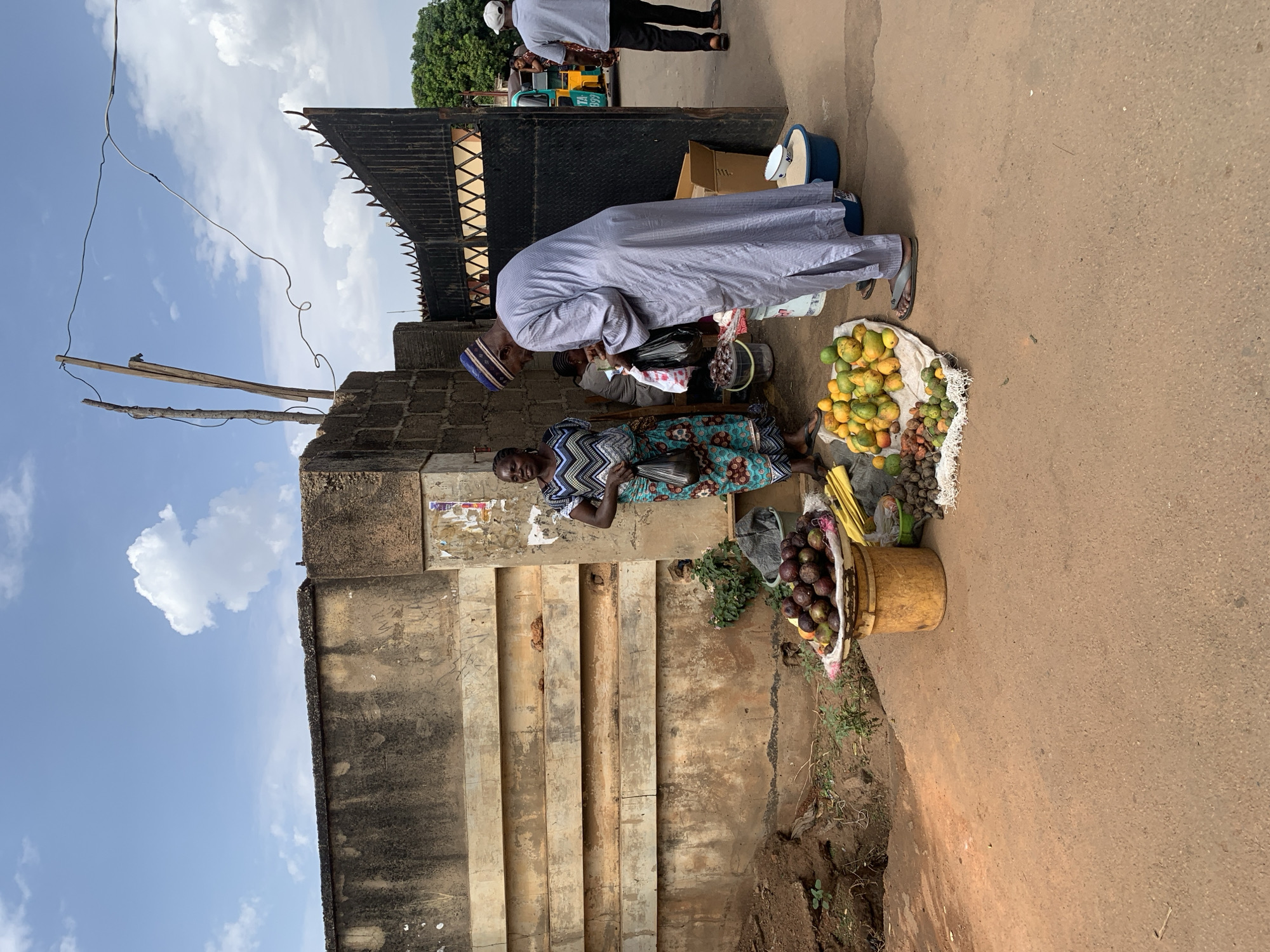 A daytime scene shows a woman seated on the ground next to a wall, selling fruit in Jos, Nigeria. She wears a patterned dress and a man in a long white robe and cap bends over the fruit to purchase some. The fruit includes mangoes, bananas, and other local varieties, displayed on a white cloth and in a bucket. Behind the man is a black metal gate. Other people are visible in the background, including someone near a yellow and green vehicle. The wall behind the woman is aged and stained, and there are telephone wires stretching overhead. The sky is partly cloudy.