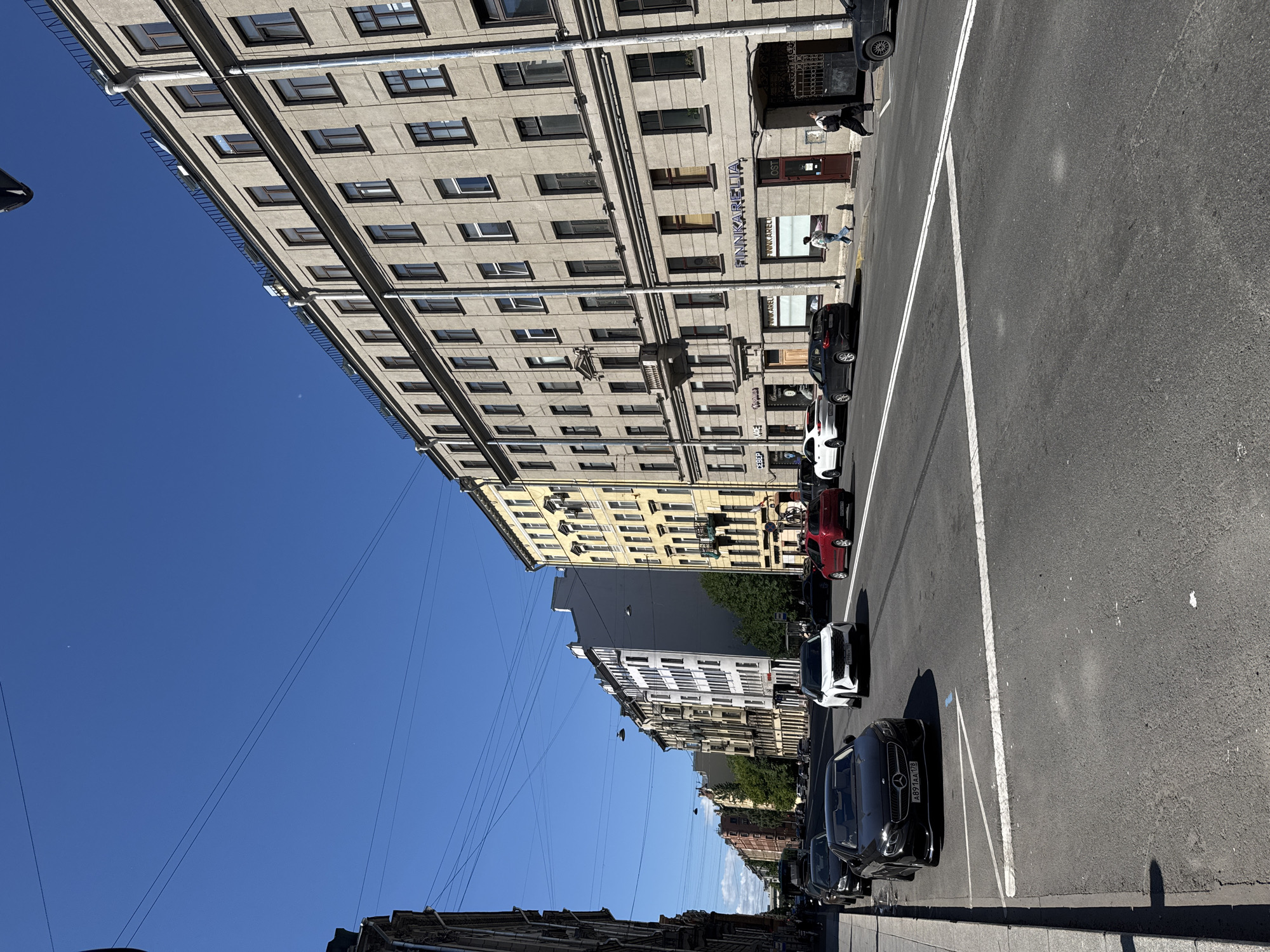 The image depicts a city street scene with tall buildings lining one side of the road. Several cars are parked along the curb, and a few people are visible walking or standing on the sidewalk. The sky is a clear, vibrant blue, suggesting a sunny day. The buildings are diverse in architectural style and color, with some featuring many windows and balconies. There are power lines crisscrossing the sky.
On one of the buildings, the text "FINNKARELIA" is visible. The cars are modern, including a black Mercedes-Benz with a license plate that reads "X891AA 178". The overall impression is of a bustling urban environment on a bright, clear day. The perspective of the photo is unusual, tilted upwards and to the side, giving a unique view of the street and buildings.