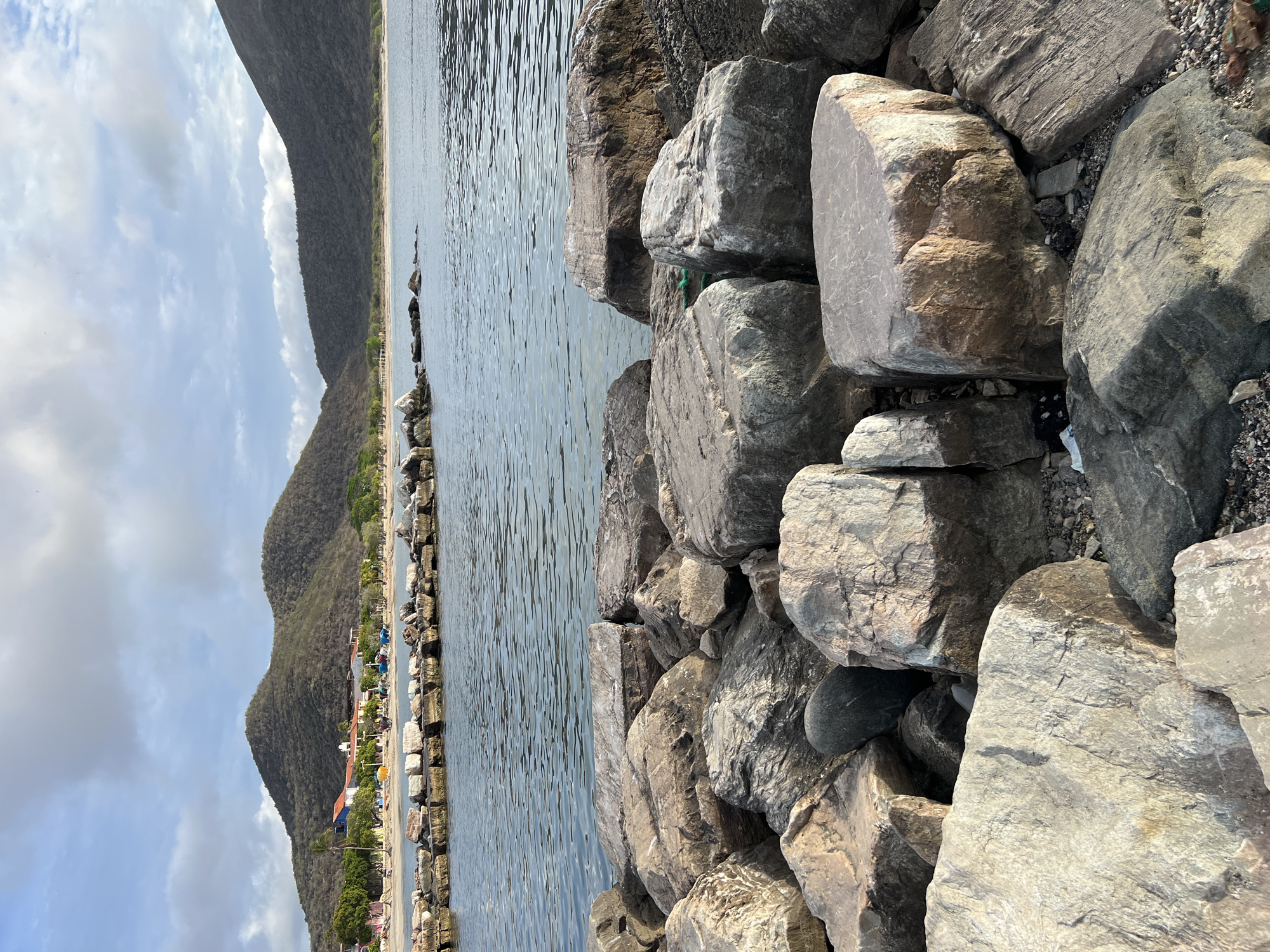 This is a photograph of the shoreline in Pedro González, Venezuela, taken from a rocky outcrop. In the foreground, large, weathered rocks are piled haphazardly. The water, a calm, grayish-blue, stretches out to the horizon, where a sandy beach is visible. Along the beach, a line of buildings, including houses and possibly shops, are nestled at the base of rolling, tree-covered hills that dominate the background. The sky above is a mix of light blue and scattered white clouds. A breakwater or jetty made of smaller stones extends into the water from the beach area. The overall scene depicts a coastal landscape with signs of habitation.