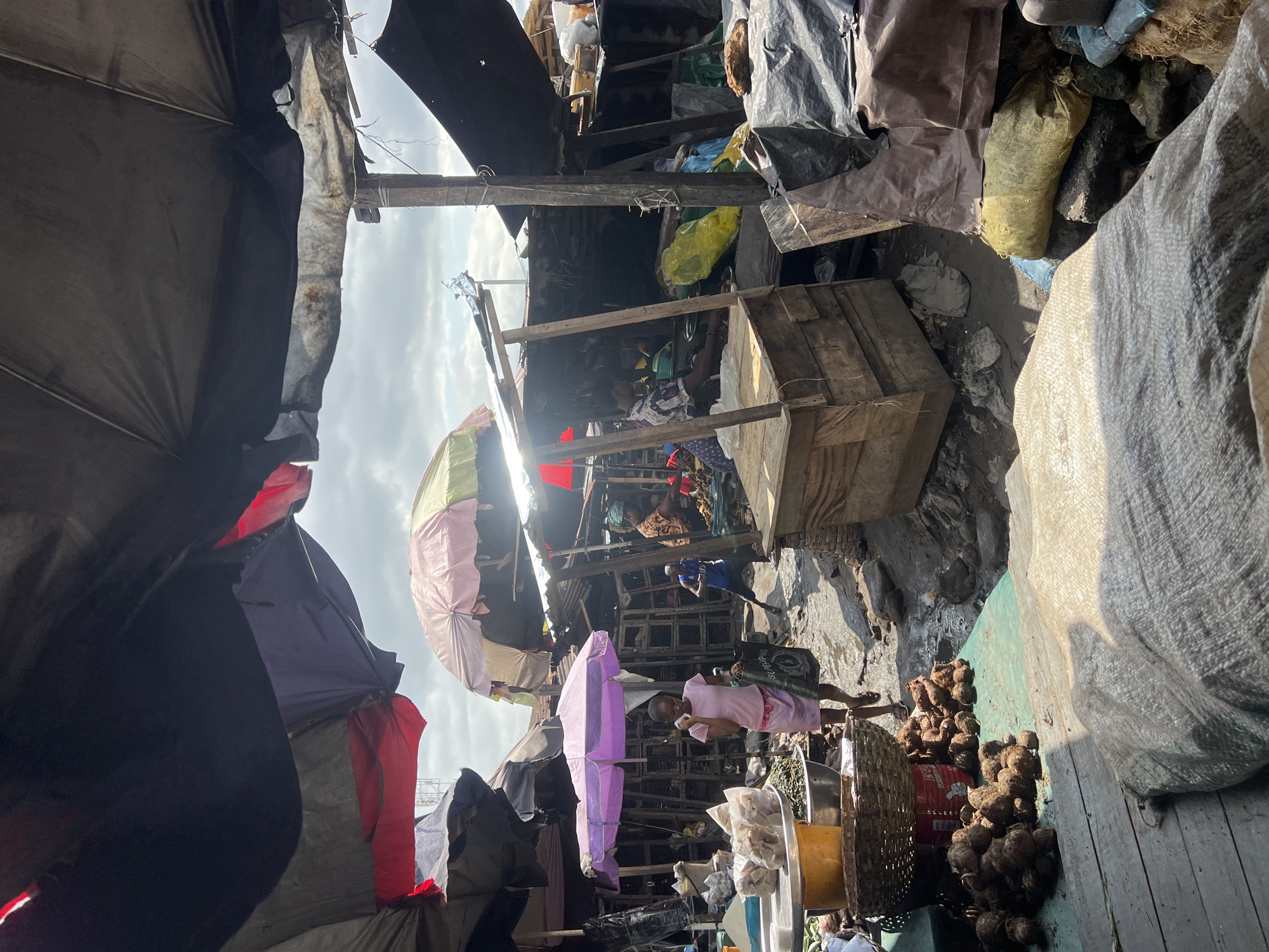 The image depicts a bustling outdoor market scene in Nkpor, Nigeria. A woman in a pink dress is walking through the market, carrying a black bag. She appears to be the central subject, with other people and market stalls surrounding her. The market is crowded with various goods, including sacks of produce, baskets, and what appear to be root vegetables piled on wooden planks. The setting is characterized by makeshift stalls constructed from wood and covered with tarpaulins and fabric, creating a somewhat disheveled but vibrant atmosphere.
Overhead, umbrellas in shades of pink, purple, and yellow provide shade, interspersed with the weathered wooden structures of the market. Sunlight streams through the gaps, suggesting it is daytime, likely late morning or afternoon given the angle of the shadows. The weather appears to be clear with some scattered clouds visible in the sky. The overall impression is one of lively commerce and everyday activity in a busy Nigerian market. The ground appears to be rough, with debris and dirt visible between the stalls. The image is oriented vertically, making it appear as if the viewer is looking upwards into the market.