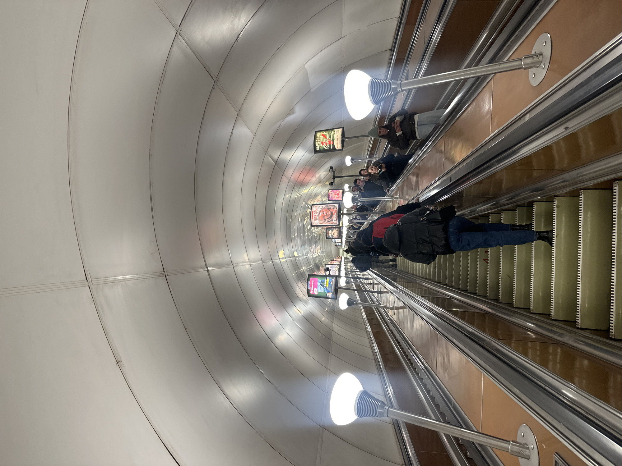 The image shows an escalator in the Saint Petersburg Metro, Russia. People are riding the escalator in both directions. There are advertisements along the walls, and distinctive globe-shaped lights illuminate the tunnel. The ceiling is constructed of curved metal panels. The escalator has silver rails and light green steps.
