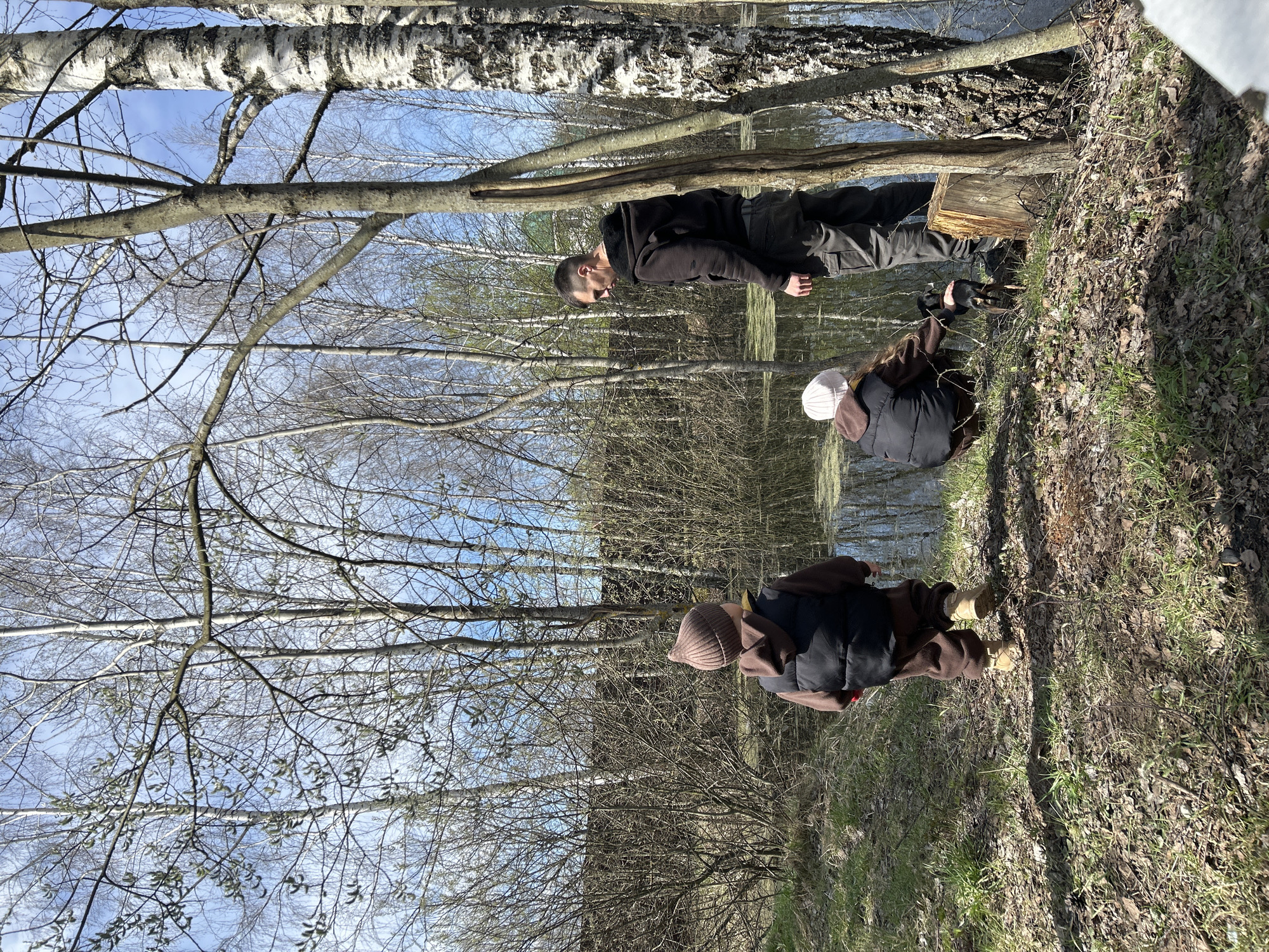 The image depicts a group of three individuals in a wooded area, likely near a body of water, possibly in the Мещёры region of Russia. The vegetation suggests an early spring setting. One individual is standing, wearing dark clothing, next to a tree stump. The second person, wearing a light-colored hat and dark jacket, is crouched down. A third person is standing closer to the foreground with a brown hat and clothing. The environment includes bare trees, sparse undergrowth, and ground covered in leaves and grass. The background suggests a body of water with reflections of trees.