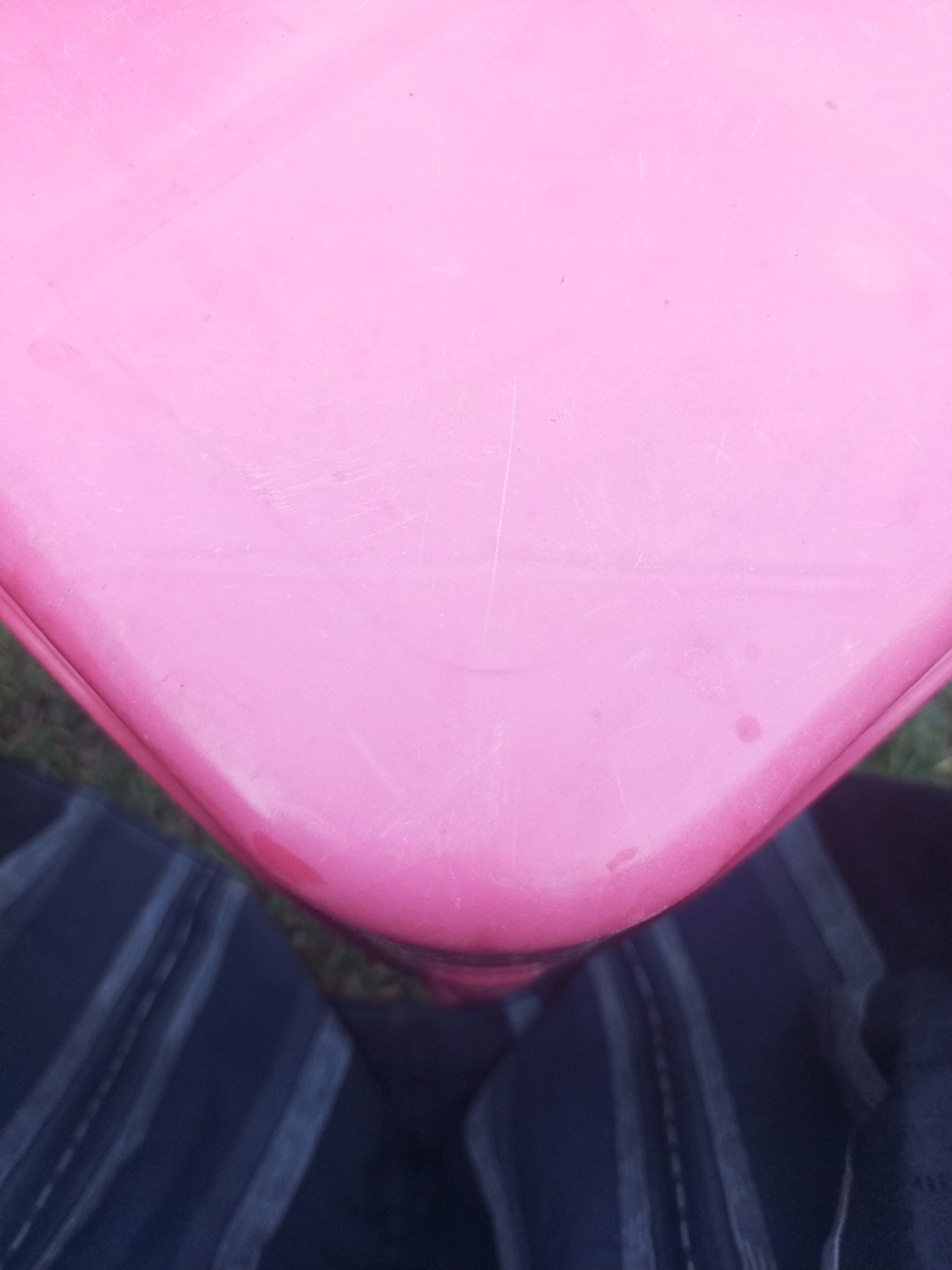 The image shows a close-up, overhead view of a curved, bright pink surface, possibly a plastic table or chair, with faint scratches and a few small, reddish-brown stains. The bottom portion of the image shows a person's dark blue and white striped pants. The background appears to be grass. There is no specific indication of the location being Lhokseumawe, Indonesia. The scene depicts a static arrangement of objects.