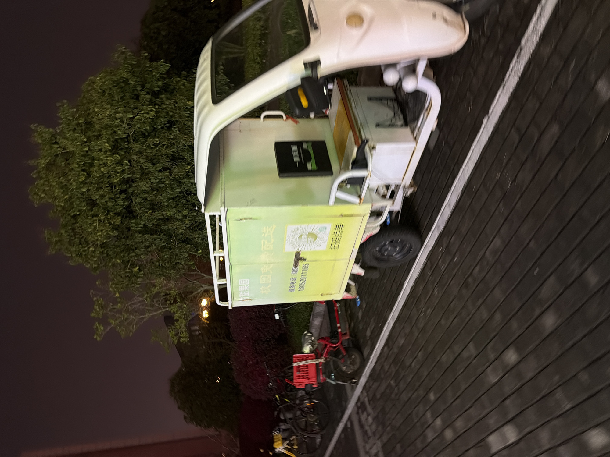 A utility vehicle sits on a paved area in Jiangning District, China, at night. Vegetation is visible in the background and several bicycles are parked close to the utility vehicle. The pavement is dark and lined.