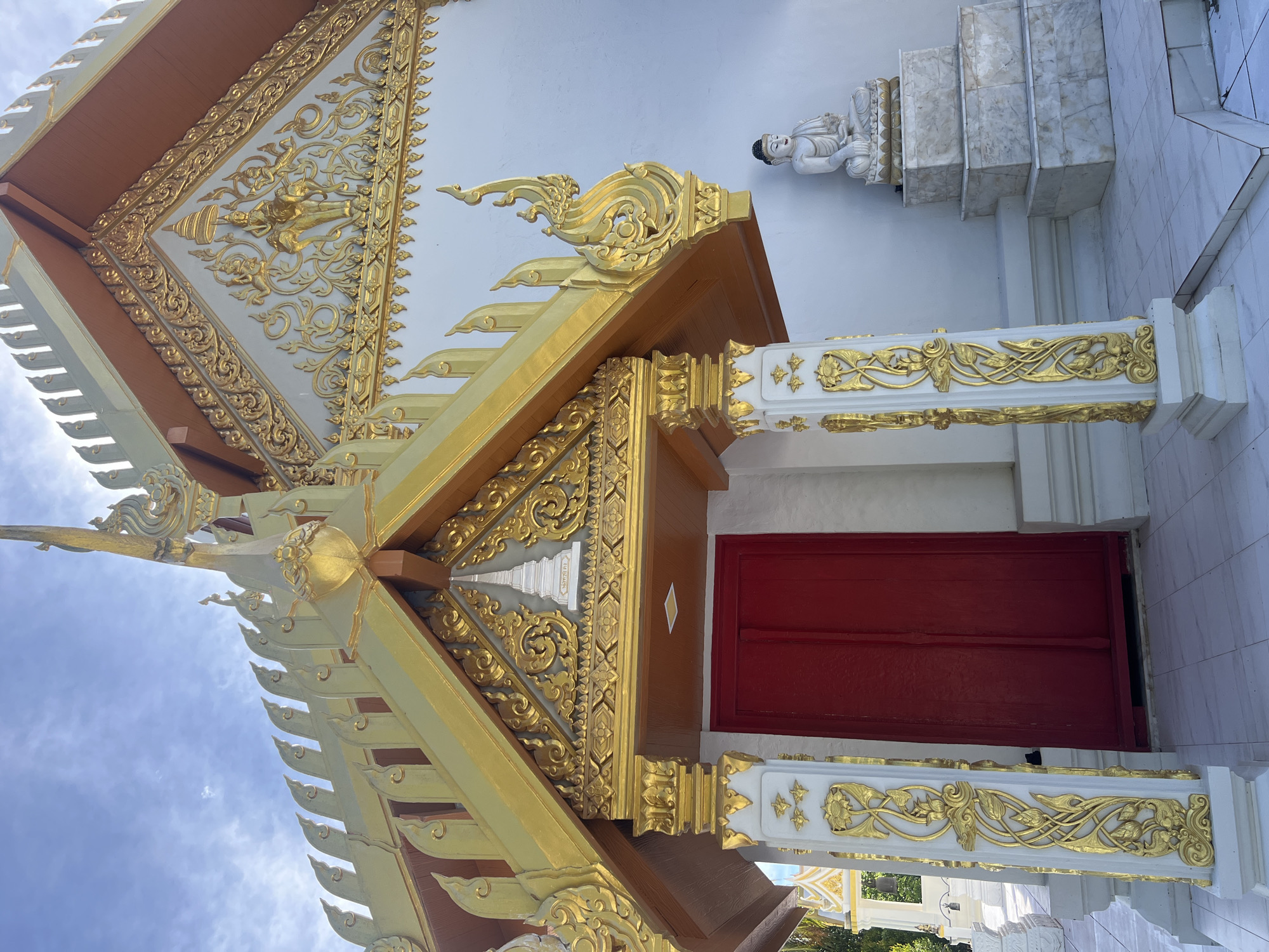 This is a vertically oriented, eye-level photograph of an ornate architectural detail of a building, likely a temple or a place of worship, in Koh Phangan, Thailand. The building features intricate golden embellishments against a white and light brown structure. A prominent part of the image is the detailed golden relief work on the eaves and columns, showcasing floral and mythical motifs. The roof structure is multi-layered with decorative elements.
To the right of the main structure, a white marble staircase leads up to a seated Buddha statue, rendered in white stone, which is placed on a pedestal. The Buddha is depicted in a meditative pose. Below the golden decorative pillars, a striking red door is visible, contrasting with the predominantly white and gold facade. The building's white walls are tiled with square, light-colored tiles on the right side of the frame.
The sky above is a pale blue with scattered white clouds, suggesting it is daytime with fair weather. The sunlight creates highlights on the golden ornamentation, emphasizing its richness and craftsmanship. The overall impression is one of grandeur, serenity, and cultural significance, typical of religious sites in Thailand. No visible text is present in the image.