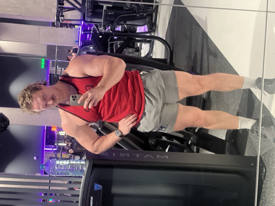 A muscular man poses in front of a gym mirror. He wears a red tank top, gray shorts, white socks, and sneakers. He holds a smartphone in his hand, taking a selfie. His face is turned slightly towards the camera. The mirror reflects gym equipment, including a Matrix treadmill. A water bottle sits on the treadmill console. Other gym-goers are faintly visible in the background. The lighting is bright and the setting is indoors.