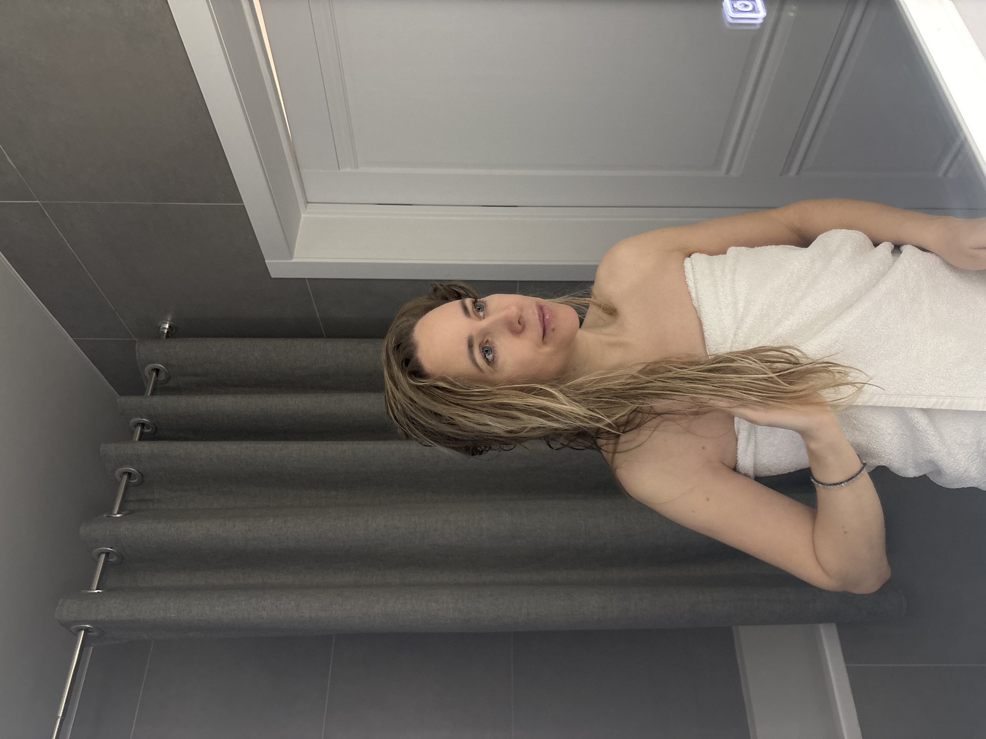 A woman wrapped in a white towel is posing in what appears to be a bathroom. The location is unknown but based on the request it could potentially be Chaikovsky, Russia. The bathroom has gray tiled walls and a gray fabric shade. There's a white-framed mirror or cabinet above her. She has blonde hair and is wearing a bracelet.