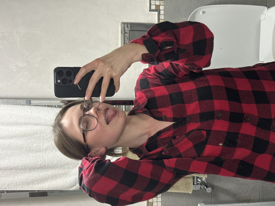 The image depicts an adult female subject taking a mirror selfie inside a bathroom. The specific location within Cava de' Tirreni, Italy, is not discernible from the visual information provided.
The subject is positioned centrally, wearing a red and black plaid flannel shirt. She has light brown hair, appears to be wearing round-framed eyeglasses, and is looking towards the camera (reflected in the mirror). Her right eye is closed in a wink, and her tongue is extended from her mouth. Her left hand is raised and positioned behind her head, while her right hand holds a dark-colored smartphone, featuring a multi-lens camera array and a circular accessory on its back, to capture the image.
The background elements indicate a bathroom environment. To the left of the subject, a chrome heated towel rail with a white towel draped over it is visible, along with a beige towel hanging below. The wall behind the subject features white textured tiles on the upper half, with a decorative border of smaller, square tiles, likely beige or light gray, separating it from a darker tiled lower section. To the right, a white toilet with its lid closed is partially visible. An electrical outlet or wall fixture is present on the tiled wall above the toilet. The lighting is uniform, suggesting an indoor setting.