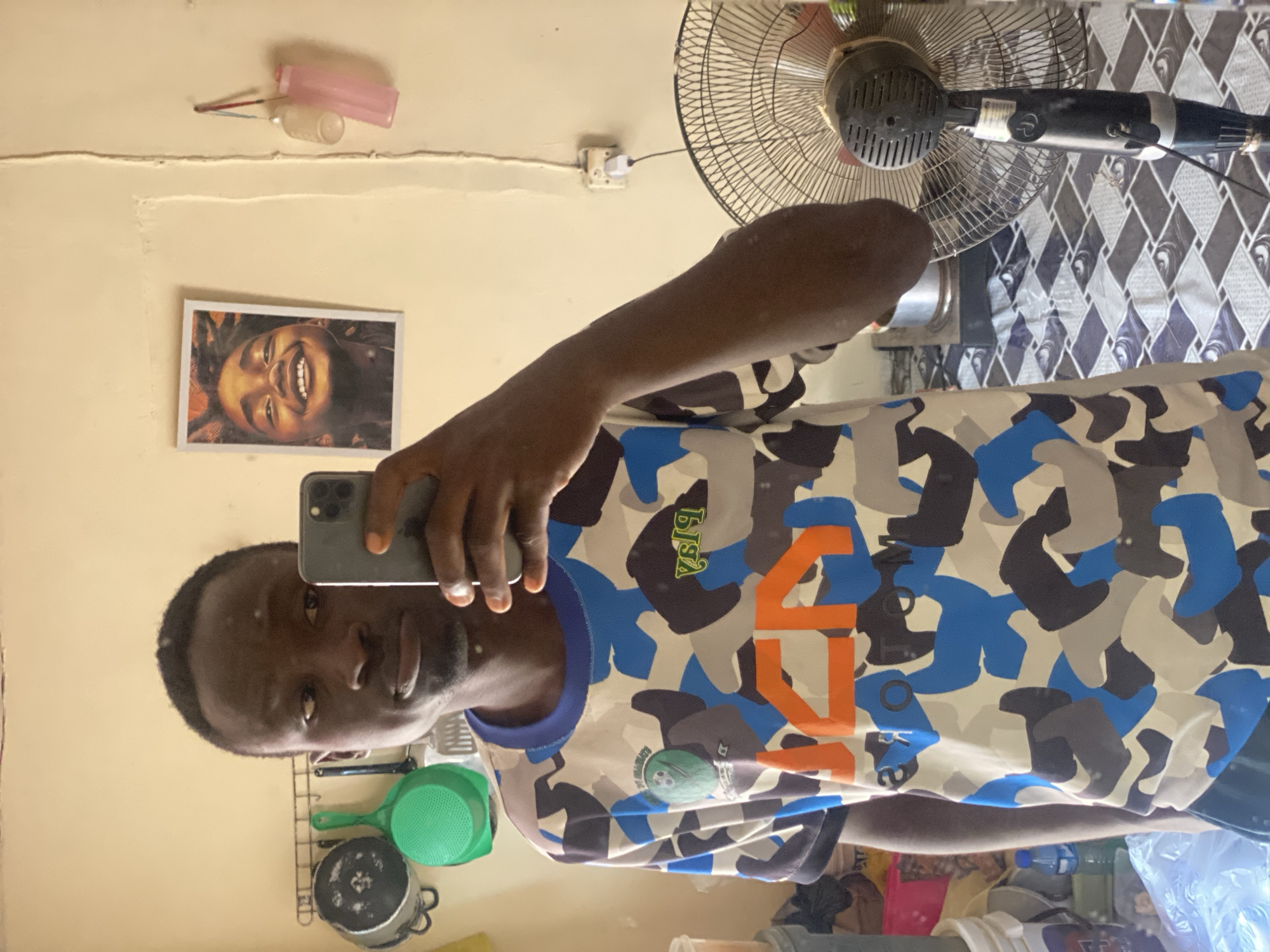 In Barden Yana, Nigeria, a young man stands in front of a mirror, taking a selfie with his phone. He is wearing a camouflage t-shirt with blue, gray, and white patterns and orange lettering on the front. The background reveals a cluttered room. On the wall behind him, a framed picture of a smiling child hangs. To the left, kitchen utensils, including pots and a green colander, are mounted on a rack. To the right, a black standing fan is visible. The room appears to be a living space with a patterned rug on the floor.