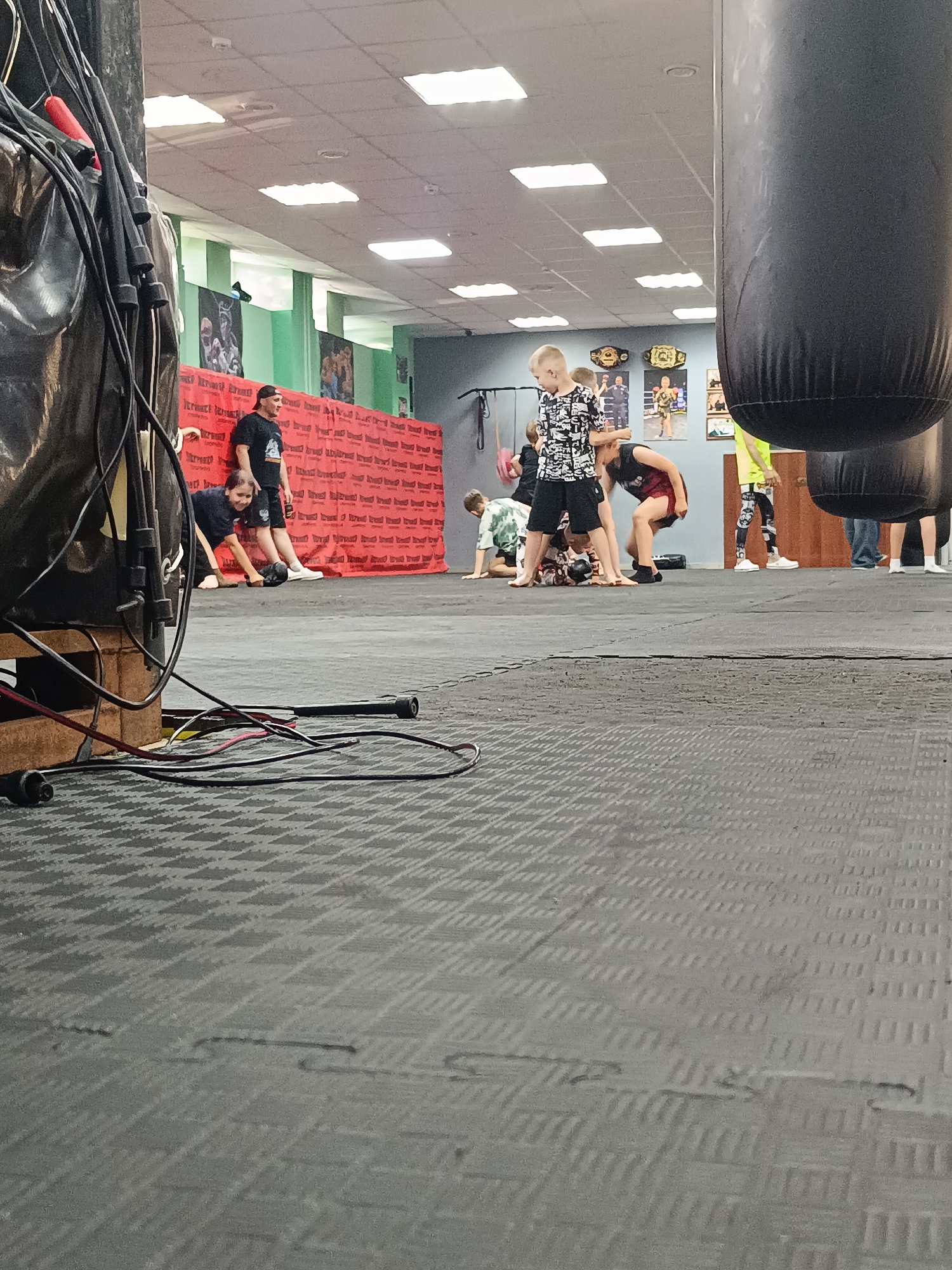 This image captures an indoor scene within a martial arts or combat sports gym, likely located in Naberezhnye Chelny, Russia. The setting features a large area covered with dark grey interlocking foam mats, typical for training. The foreground on the left shows a dense cluster of black cables, wires, and several jump ropes hanging over a dark, possibly protective, cover, with a wooden pallet or box beneath. On the far right foreground, part of a large black punching bag is visible.
In the mid-ground, several children and at least two adults are present. A young boy in a black and white patterned shirt stands centrally, looking down. Around him, other children are engaged in various movements on the mat, some bending over, one appearing to be on their knees or the ground, suggesting warm-ups, drills, or grappling practice. An adult male in a black t-shirt and shorts, wearing a backward baseball cap, is visible in the background leaning against a red wall covering, observing the children. Another adult is partially visible in the far background right.
The background wall is adorned with a red panel featuring repeated text that appears to be part of a brand or gym name. Above this, the wall transitions to a light green section. Several framed posters depicting fighters or martial arts scenes, along with two large, ornate championship belts, decorate the upper part of the wall. The ceiling is illuminated by numerous square fluorescent light panels. The overall atmosphere suggests an active training session in a dedicated facility.