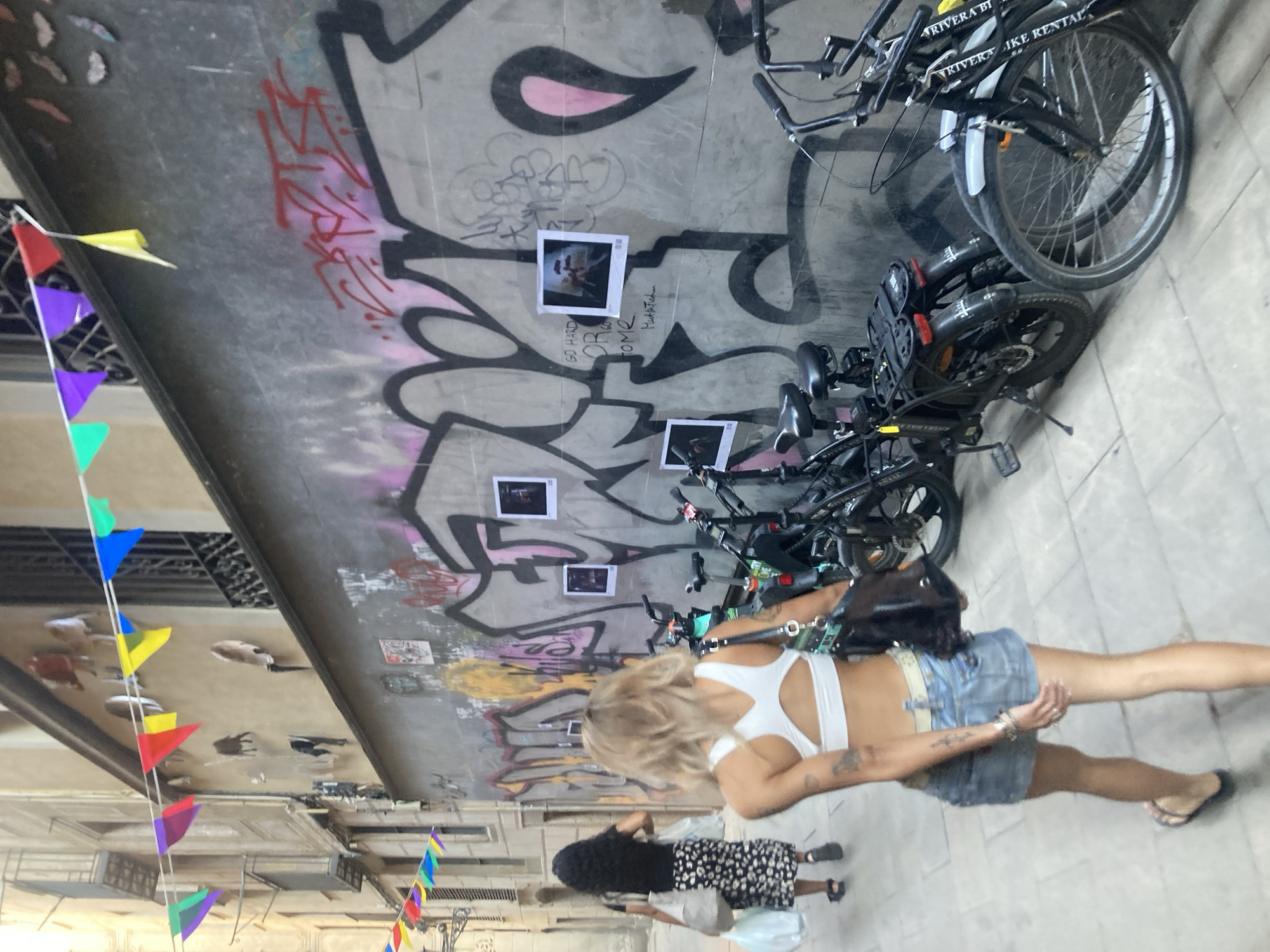 The image captures an outdoor scene, likely in Barcelona, Spain, with a focus on a wall covered in graffiti. In the foreground, a woman with blonde hair and a white top and denim shorts is walking away from the camera. Behind her, another person with dark curly hair is also visible. The wall behind them is adorned with vibrant graffiti art, including bold black and pink designs. Several small framed photographs are attached to the graffiti-covered wall.
To the right of the wall, a row of parked bicycles is visible, some with "RIVERA BIKE RENTAL" printed on them. String lights with colorful triangular flags are draped across the scene, suggesting a festive atmosphere or an event. The overall lighting and shadows indicate it might be daytime. The setting appears to be a street or alleyway with buildings and windows visible in the background, contributing to the urban feel.