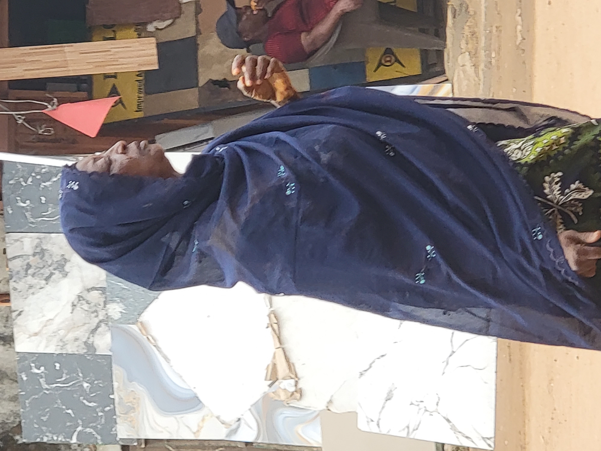 An elderly woman wearing a dark blue head covering decorated with teal embroidery sits in Maiburiji, Nigeria. She is on or adjacent to a tiled surface comprised of square sections with a gray and white marbleized pattern and solid white marbled sections. She is wearing a green and black patterned garment under the head covering. Her hand is raised near her face. Another person wearing a red shirt is blurry in the background. Miscellaneous objects including boards and a red flag are also visible in the background. The light indicates an outdoor setting during the daytime.