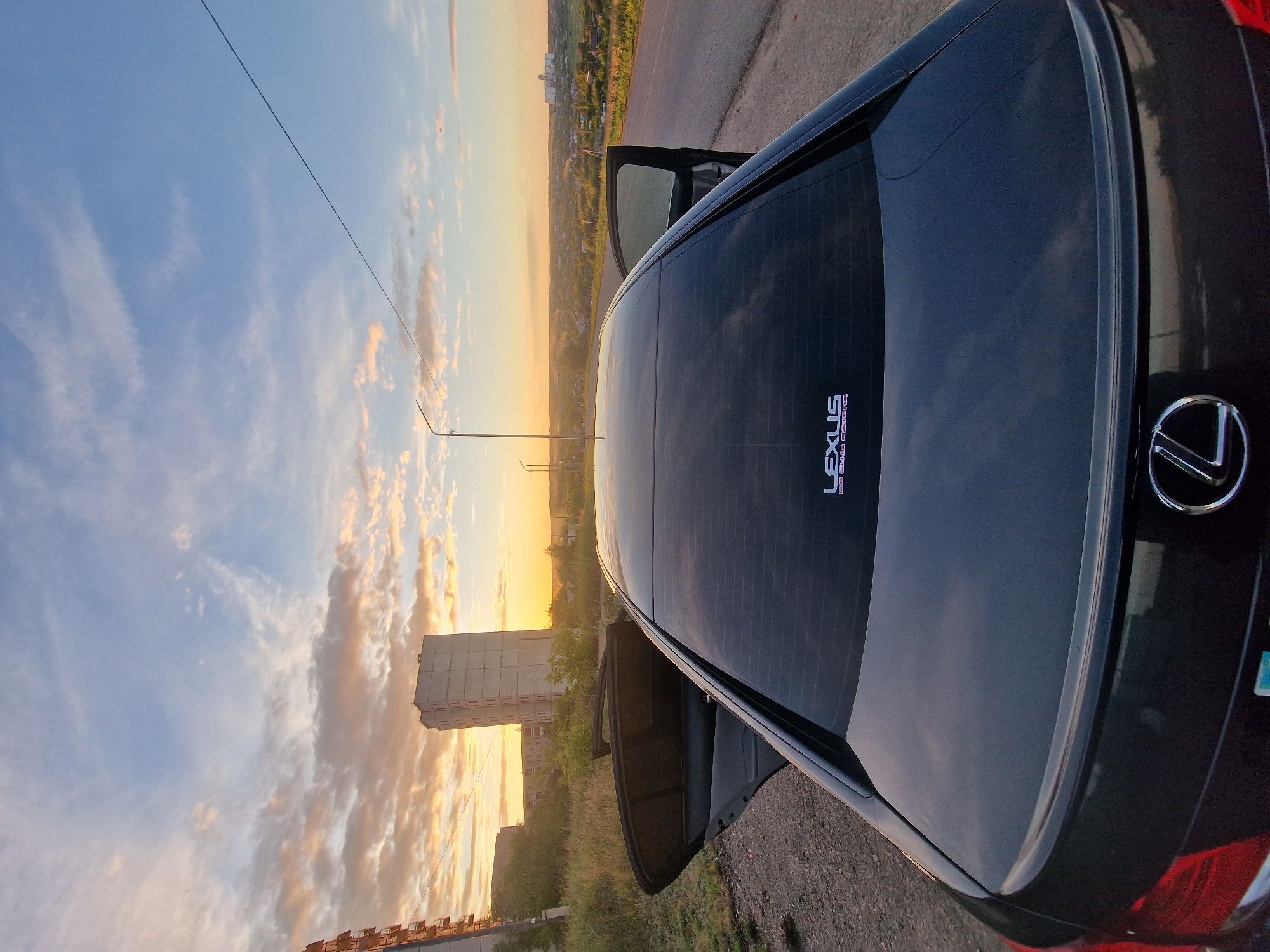 The image shows the rear of a black Lexus sedan parked on the side of a road at dusk in Kashgar City, China. The sky is filled with colorful clouds, illuminated by the setting sun, casting a warm glow over the landscape. In the background, there are buildings and trees, with a prominent tall, concrete building on the left. The car's rear window is tinted, and a "LEXUS" logo is visible on the lower right side of the trunk. The driver's side door is open. The overall scene suggests a peaceful and serene moment during sunset.