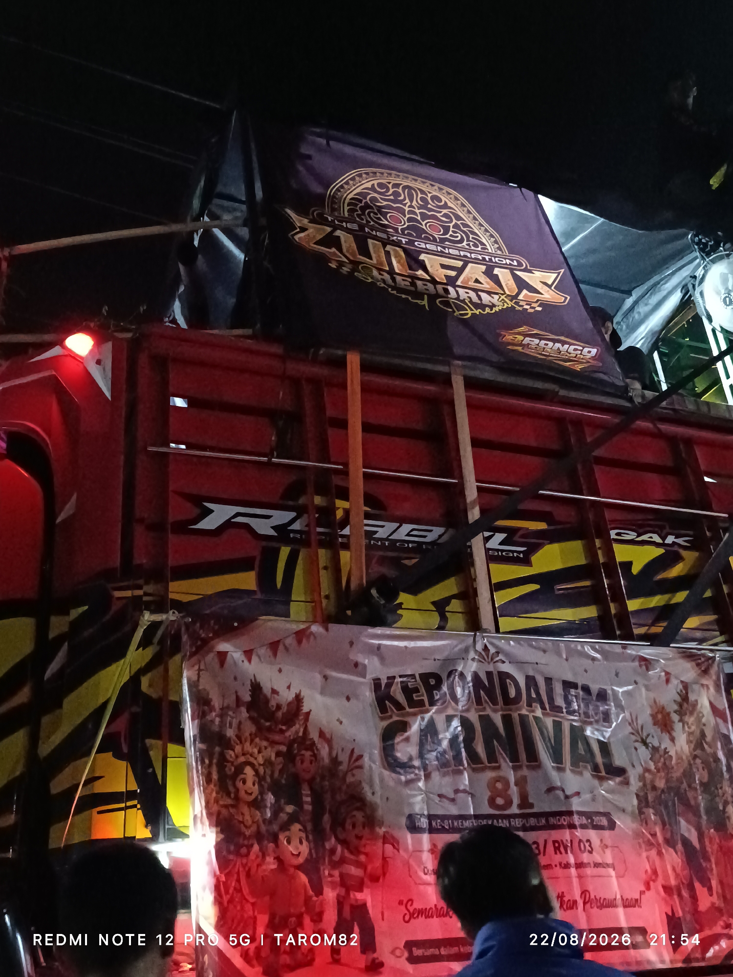 The image captures a night scene featuring a decorated truck adorned with banners and lights, likely for a local event. A large purple banner with gold lettering, reading "ZULFAIX NEXT GENERATION SHEBORN SOUND SYSTEM BRONCO", is prominently displayed at the top. Below this, a red truck with yellow and black graphics is visible. A white banner with festive imagery and text, including "KEBONDALEM CARNIVAL 81" and "HD KE-81 KEMERDEKAAN REPUBLIK INDONESIA · 2026", is hung across the front of the truck, illuminated by red lights. The lower part of the banner contains additional text in smaller font. The timestamp "22/08/2026 21:54" and "REDMI NOTE 12 PRO 5G | TAROM82" are overlaid on the image. The location is identified as the city of Mundusewu, Indonesia. Several individuals are partially visible in the foreground, with their backs to the camera. The environment appears to be outdoors, possibly a street or an open area, with overhead wires and ambient lighting contributing to the nighttime atmosphere. The overall impression is that of a celebratory or promotional event.