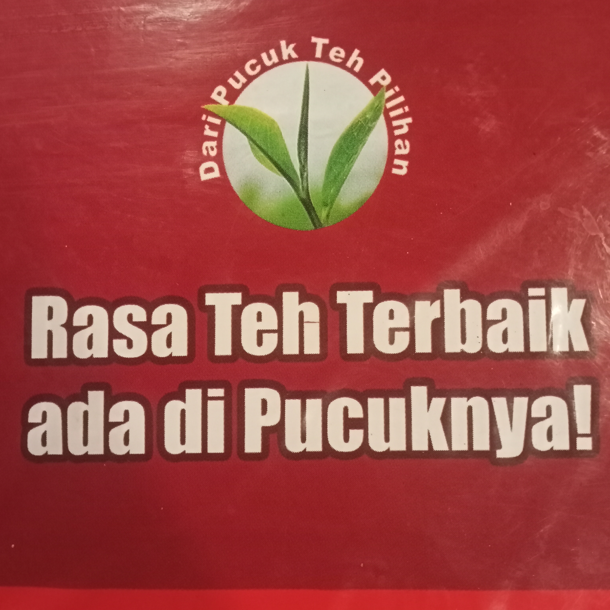 The image is a close-up of a red background with text and a circular graphic. The circular graphic, located at the top center, features a green depiction of tea leaves and a sprout against a white background with a subtle grid pattern. Encircling this graphic in white text is "Dari Pucuk Teh Pilihan," which translates to "From the Top of Chosen Tea."
Below the graphic, two lines of larger white text with a darker outline read:
Line 1: "Rasa Teh Terbaik" (The Best Tea Flavor)
Line 2: "ada di Pucuknya!" (is in its Tip!)
The overall presentation suggests branding or advertising for a tea product, emphasizing the quality derived from the tea's bud or tip. There are no people, actions, or specific environmental details beyond the red background, which appears to be a flat surface, possibly a poster or packaging. The location in Ketowan, Indonesia, is not visually indicated in the image itself; this is external information.