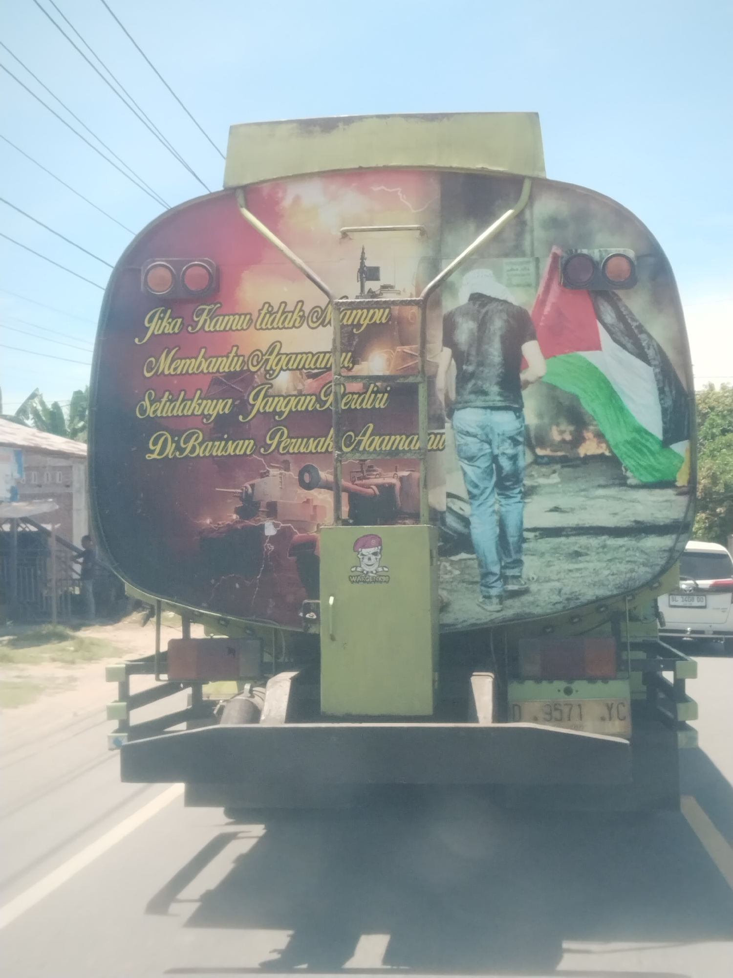 Observed on a street in Seuneubokteungoh, Indonesia, is the rear of a large, olive-green tank truck. A mural is painted on the back of the tank depicting a man from the rear wearing jeans and holding the flag of Palestine, while a tank is shown in the background. Written above the mural in gold lettering is a phrase which translates roughly to: “If you are unable to help your religion, at least don’t stand with those who destroy it.” A small green utility cabinet affixed to the rear of the truck features a sticker. The license plate of the truck is labeled “D 3571 YC.” A white vehicle is visible in the background.