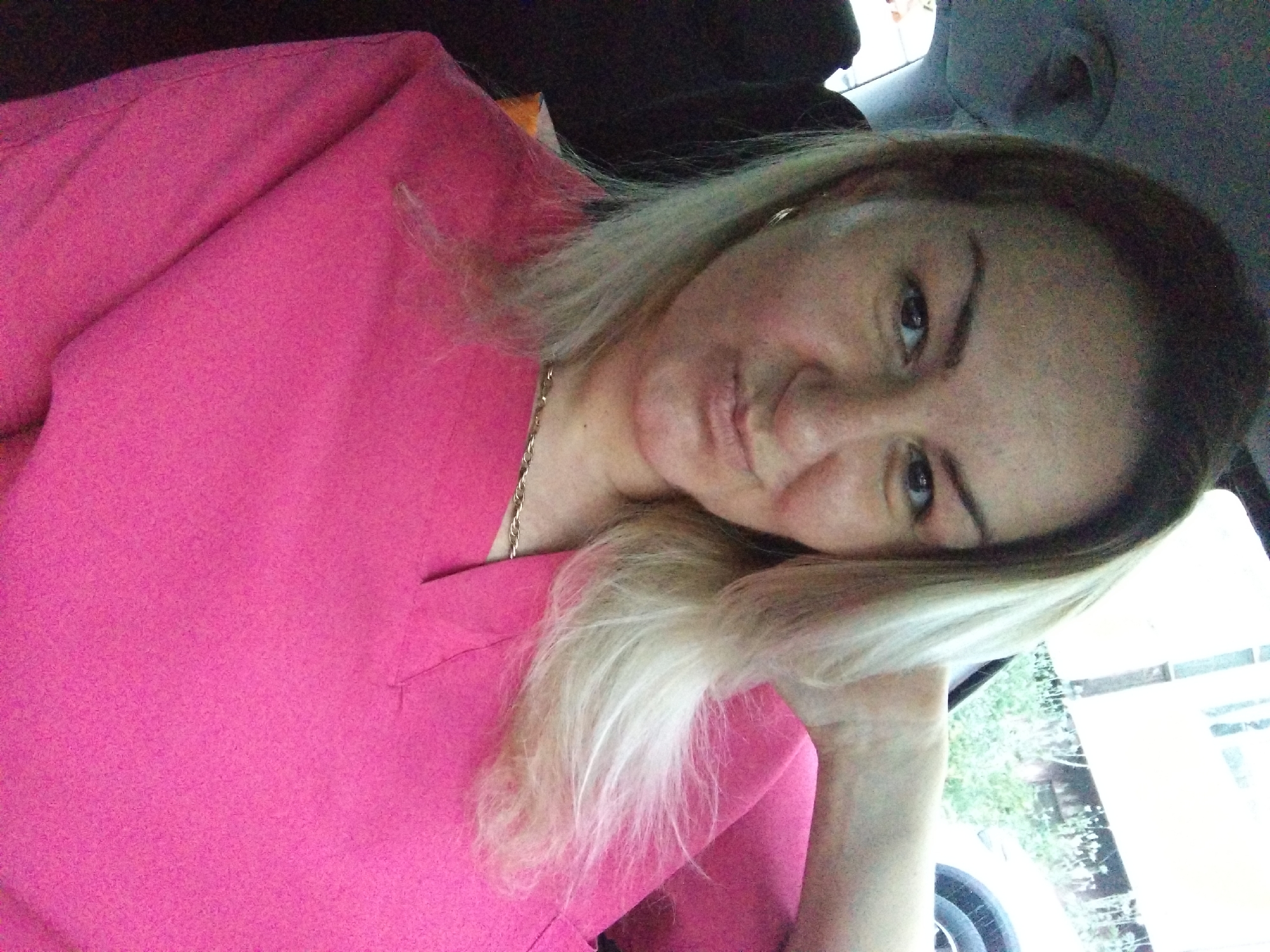 A woman with light brown hair, wearing a pink shirt, is in what appears to be a car. Her head is tilted, with her left hand propping it up. Outside the car window, there is a blurry view of foliage and a building. There is not enough information to identify a specific location in Stara Zagora, Bulgaria.