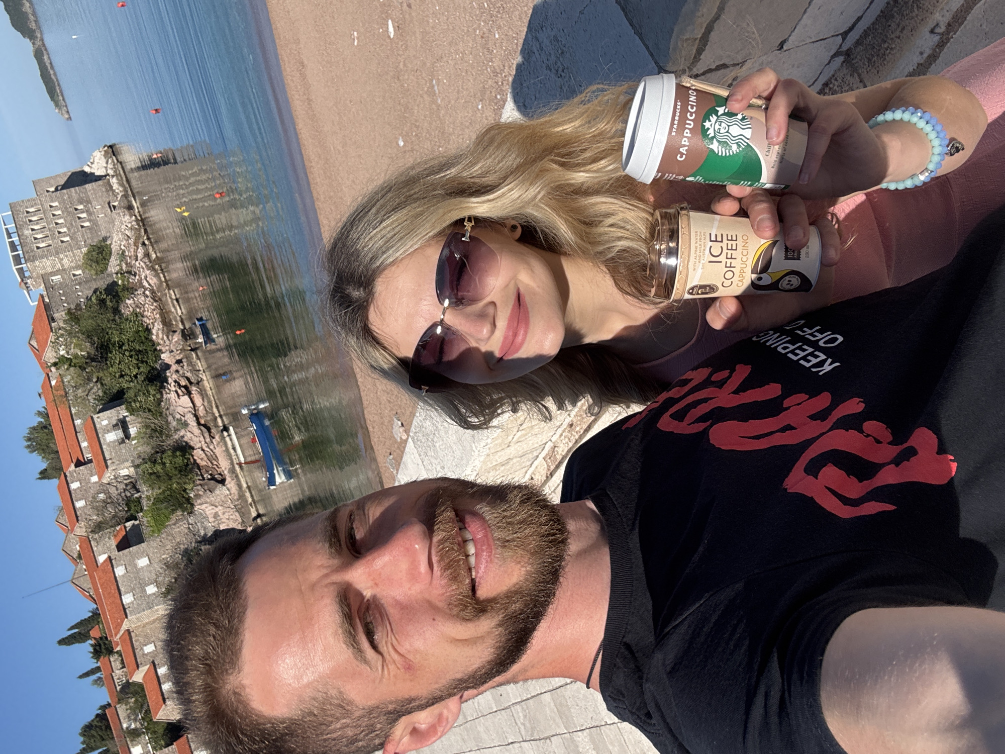 A cheerful selfie captures two individuals, a man and a woman, enjoying drinks by the coast in Sveti Stefan, Montenegro. The scene is brightly lit under a clear blue sky, suggesting a sunny day.
The woman, with long blonde hair and sunglasses, smiles broadly while holding two ready-to-drink coffee beverages. One is a Starbucks Cappuccino can, bearing the text "STARBUCKS CAPPUCCINO," "Chill," and "Bold espres." The other is a bottle of "ICE COFFEE CAPPUCCINO" featuring a penguin illustration, with visible text including "WITH ALPINE WATER," "MILK FROM THE AUSTRIAN ALPS," and "230ml." She wears a light-colored, beaded bracelet on her wrist.
Beside her, a man with a beard smiles at the camera. He is wearing a black t-shirt with red lettering, part of which reads "KEEPING OFF."
The background reveals the picturesque coastal town of Sveti Stefan, characterized by its distinctive stone buildings with red-tiled roofs, situated on a rocky islet. These structures are reflected in the calm, blue waters of the Adriatic Sea. A sandy beach lines the shore, and a few small boats are visible in the water. Both individuals appear relaxed and happy, enjoying their time by the sea.