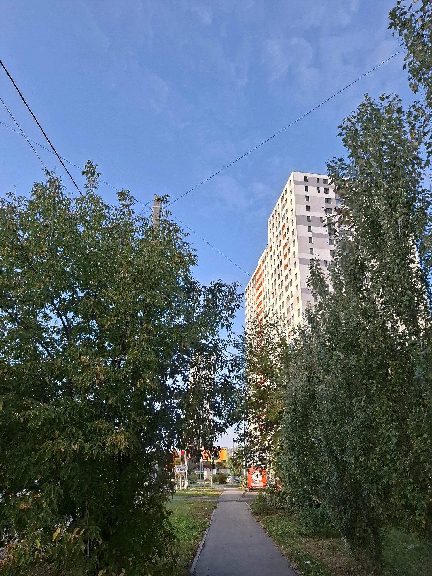 The image depicts a residential area in Tyumen, Russia, featuring a tall, modern apartment building with a facade of white, grey, and orange elements. The building is situated behind a row of lush green trees, which partially obscure its lower levels. In the foreground, a paved pedestrian walkway runs alongside a grassy area and more trees. Overhead, power lines crisscross the clear blue sky, which is dotted with faint clouds. Signs of urban infrastructure, such as streetlights and traffic signals, are visible in the distance. The scene conveys a sense of everyday urban life under a bright sky.