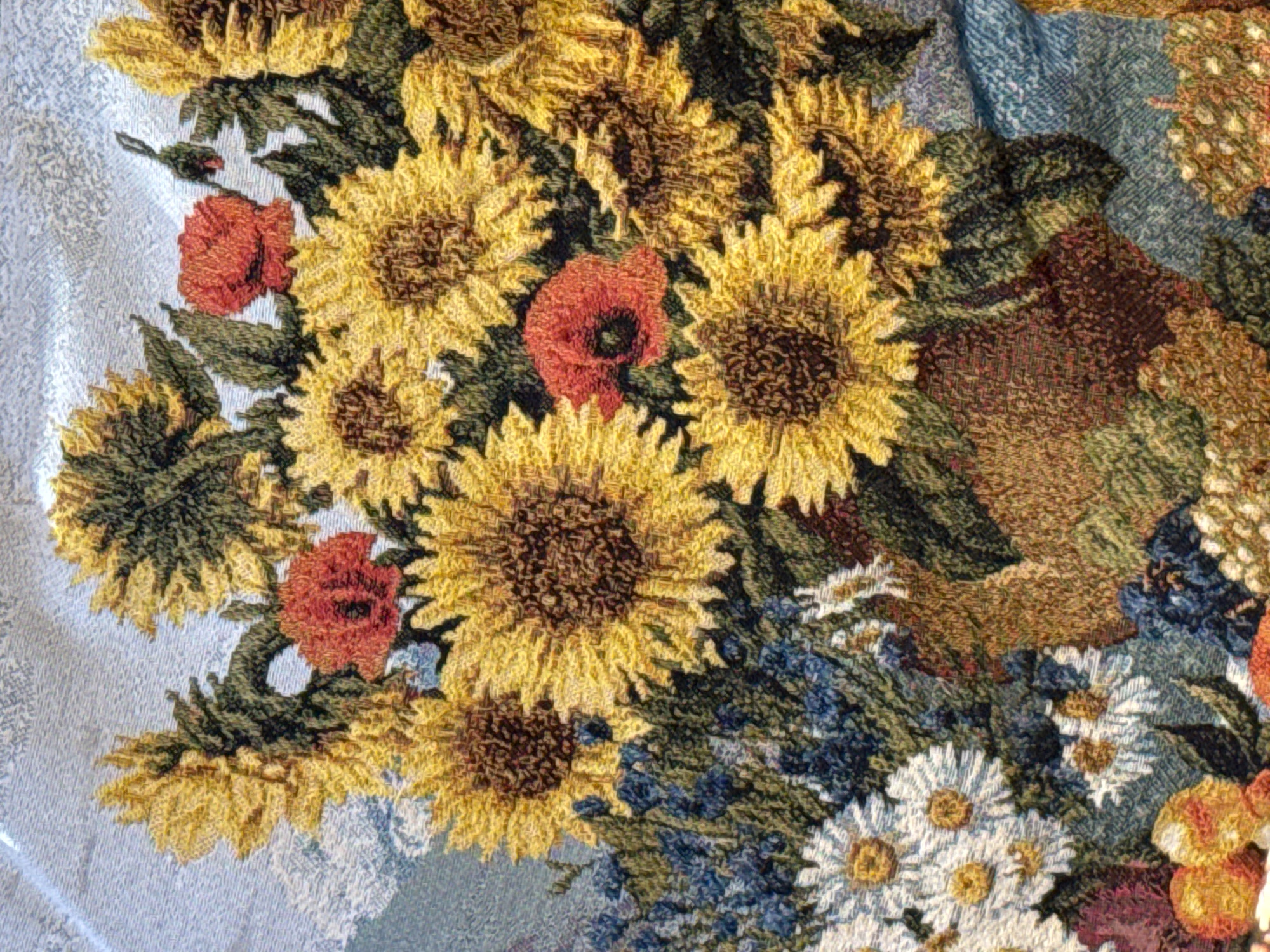 The image shows a close-up of a floral tapestry. The prominent flowers are sunflowers, rendered in shades of yellow and brown. Interspersed among the sunflowers are red poppies and white daisies. Blue and green foliage fills the spaces between the larger blooms. The background is a light blue fabric with a subtle woven pattern. The image is likely a detail shot, focusing on the texture and colors of the tapestry. There are no people, specific actions, or interactions visible in the image. The image does not show the location in the city of Ketoshi, Russia.