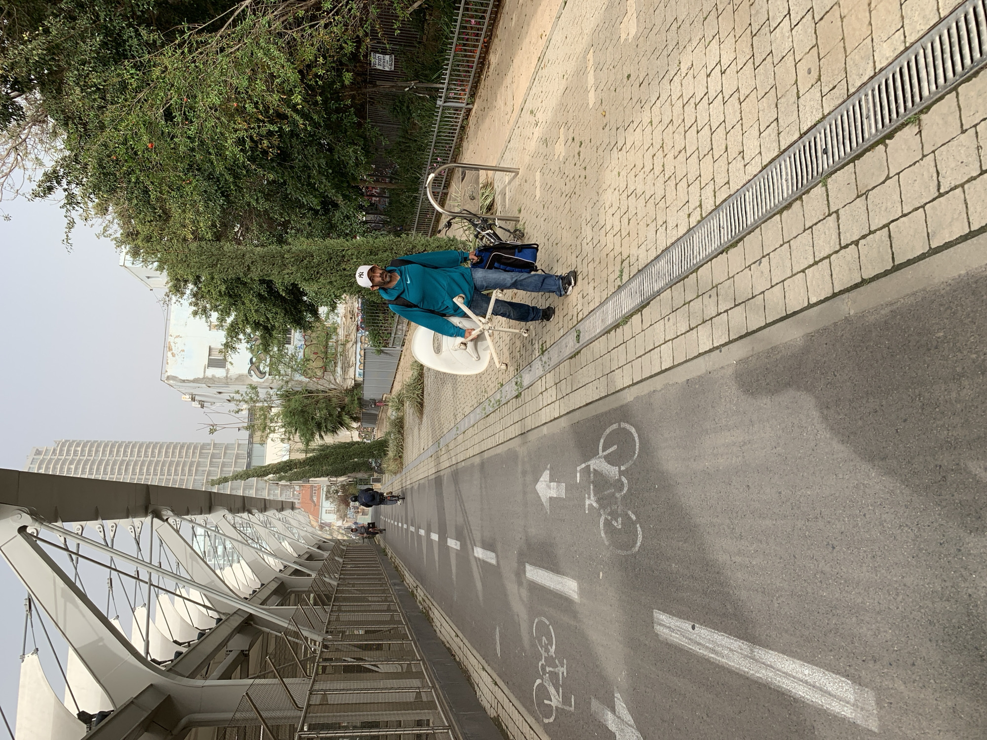 A man walks on a pedestrian and bicycle path in Tel Aviv, Israel. The path is marked with bicycle symbols and direction arrows. The man, wearing a turquoise jacket and a white baseball cap, carries a white office chair. A bicycle is parked beside him with a blue bag attached. In the background, a modern bridge structure and a high-rise building are visible. The path is bordered by vegetation and a metal fence. Two cyclists ride in the distance.