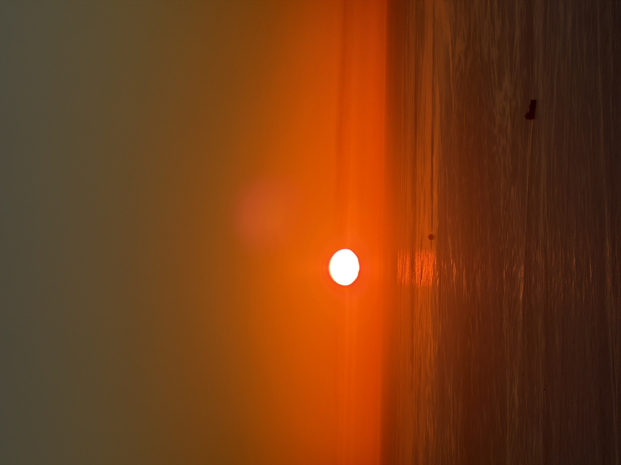The image captures a serene sunset or sunrise over a body of water, likely an ocean, given the Cape Town location context. The sun, a bright and intense white circular orb, is positioned low on the horizon, emanating a powerful glow.
The sky on the left side of the frame transitions from a muted, hazy brown-grey at the far edge to a rich, luminous orange and amber as it approaches the sun. This atmospheric gradient suggests the presence of mist, haze, or possibly dust in the air.
The right side of the image depicts the water's surface, which beautifully reflects the warm hues of the sky and sun. Numerous distinct vertical streaks of light and dark are visible across the water, indicating ripples or small waves catching and refracting the light. The water itself appears a deep, warm reddish-brown or dark orange.
Two very small, dark, and indistinct shapes can be observed on the water's surface in the distance; one appears as a subtle, rectangular form, possibly a distant boat or buoy, while the other is a tiny speck further to the right. The overall scene evokes a calm and tranquil atmosphere, characterized by the low, diffused light and the dominant warm color palette. There is no visible text in the image.