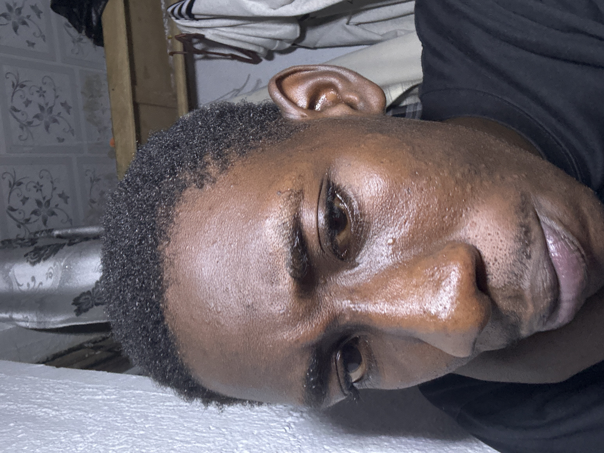 The image is a close-up, candid portrait of a Black male. He is looking down and to his left, with his head angled and slightly tilted. His skin is dark and appears slightly sweaty. His hair is short and dark. The background is out of focus but suggests an indoor environment with patterned wallpaper, a wooden shelf, and clothing hanging on a rack. A white textured wall is visible on the left side of the frame. The overall lighting is dim, casting some shadows. The specific location in the city of Durimi, Nigeria cannot be determined from the visual information provided.