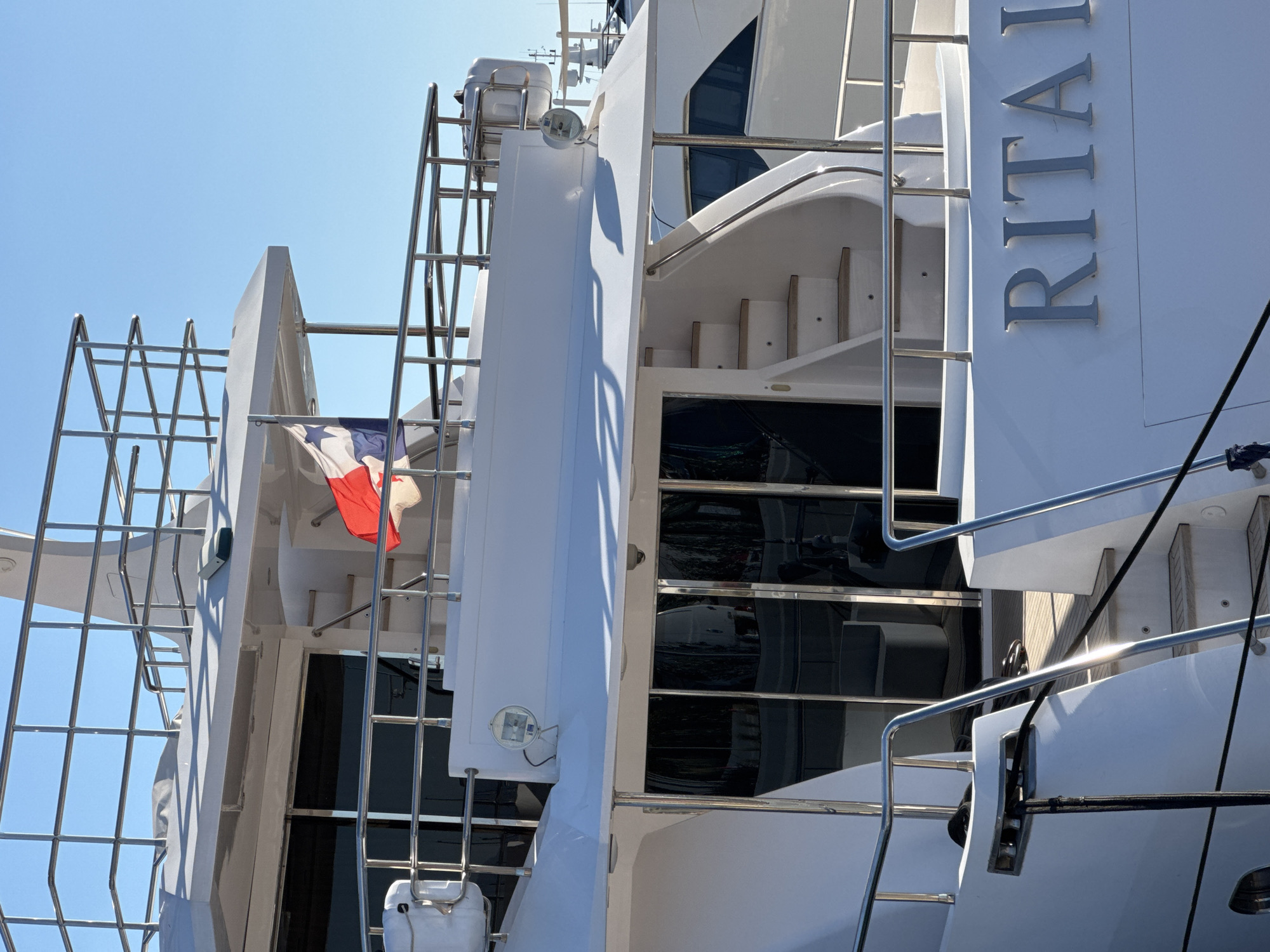This media file captures a close-up, angled view of a white luxury yacht. The prominent object is the yacht's structure, characterized by its sleek, white surfaces and polished metal railings and ladders. On the side of the yacht, the name "RITA I" is visible in large, silver, raised lettering. A small French flag is attached to a railing, fluttering slightly. The scene is set against a clear, bright blue sky, suggesting a sunny day. The lighting indicates it is daytime, likely late morning or early afternoon, with strong sunlight casting shadows. The image focuses on the yacht's architectural details and the textures of its materials, with reflections visible on the dark-tinted windows. No people are visible. The setting appears to be a marina or a harbor, possibly in Moschato, Greece, given the provided location context, with the calm water implied by the stable placement of the vessel.