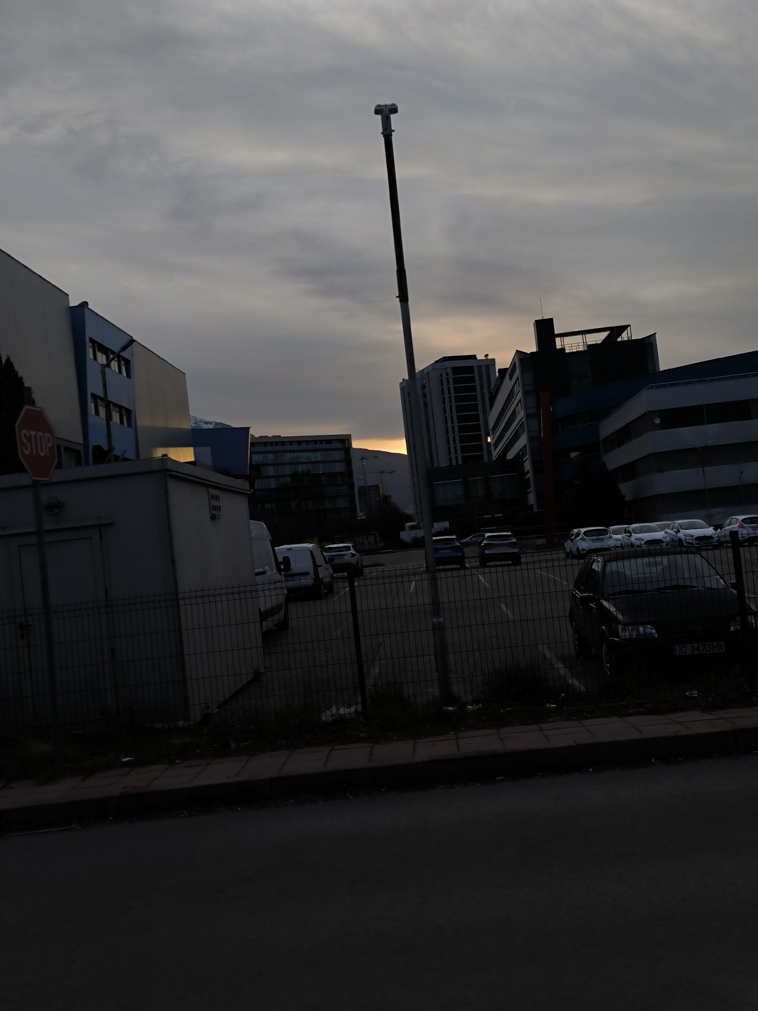 The image depicts a parking lot in Sofia, Bulgaria, at dusk. Several buildings, including a multi-story office building, surround the lot. A tall pole stands in the center. A STOP sign is visible on the left. Various vehicles are parked. A chain-link fence separates the parking lot from the road. The sky is overcast. A dark car is parked near the fence. The asphalt street in the foreground is dark. The number plate of the dark car is visible and reads "C 3473 HX". The general area appears industrial or commercial.