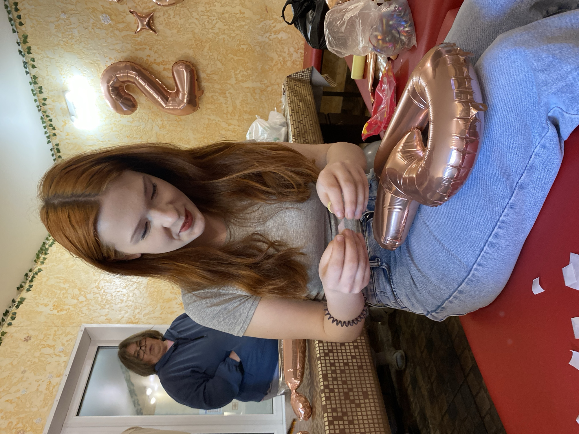A scene indoors in the city of Chaikovsky, Russia, shows a woman with red hair, wearing a gray t-shirt and jeans, seated with a partially inflated rose-gold number balloon in her lap, appearing to tie it closed. The balloon is shaped like the number "2." Another woman is visible in the background, seated near a window, wearing glasses and a blue sweater with arms crossed. A red surface is partially visible beneath the first woman. The walls are textured and patterned, and there is a partially visible decorative garland on the wall, along with additional "2023" shaped balloons. Bags and containers filled with party supplies are visible in the background.