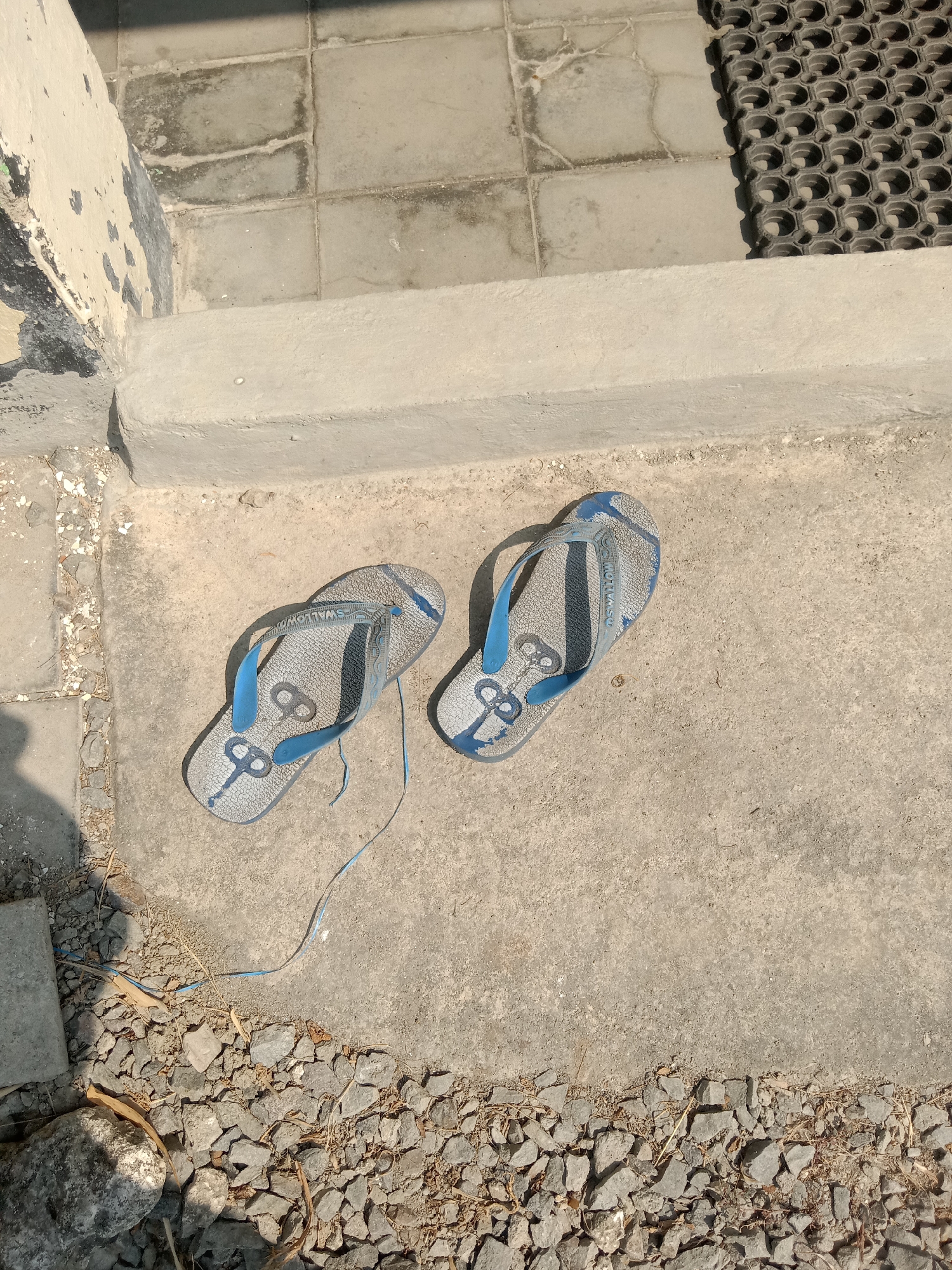 In Stiaji, Indonesia, a pair of blue and grey flip-flops lie on a concrete surface. The flip-flops are positioned side-by-side, with the left one slightly overlapping the right. A thin blue string is attached to the left flip-flop and trails onto the concrete. The ground around the flip-flops is a mixture of concrete and small stones. In the background, a tiled surface and a dark rubber mat are visible, casting shadows. The scene appears to be outdoors, possibly near an entrance or pathway.