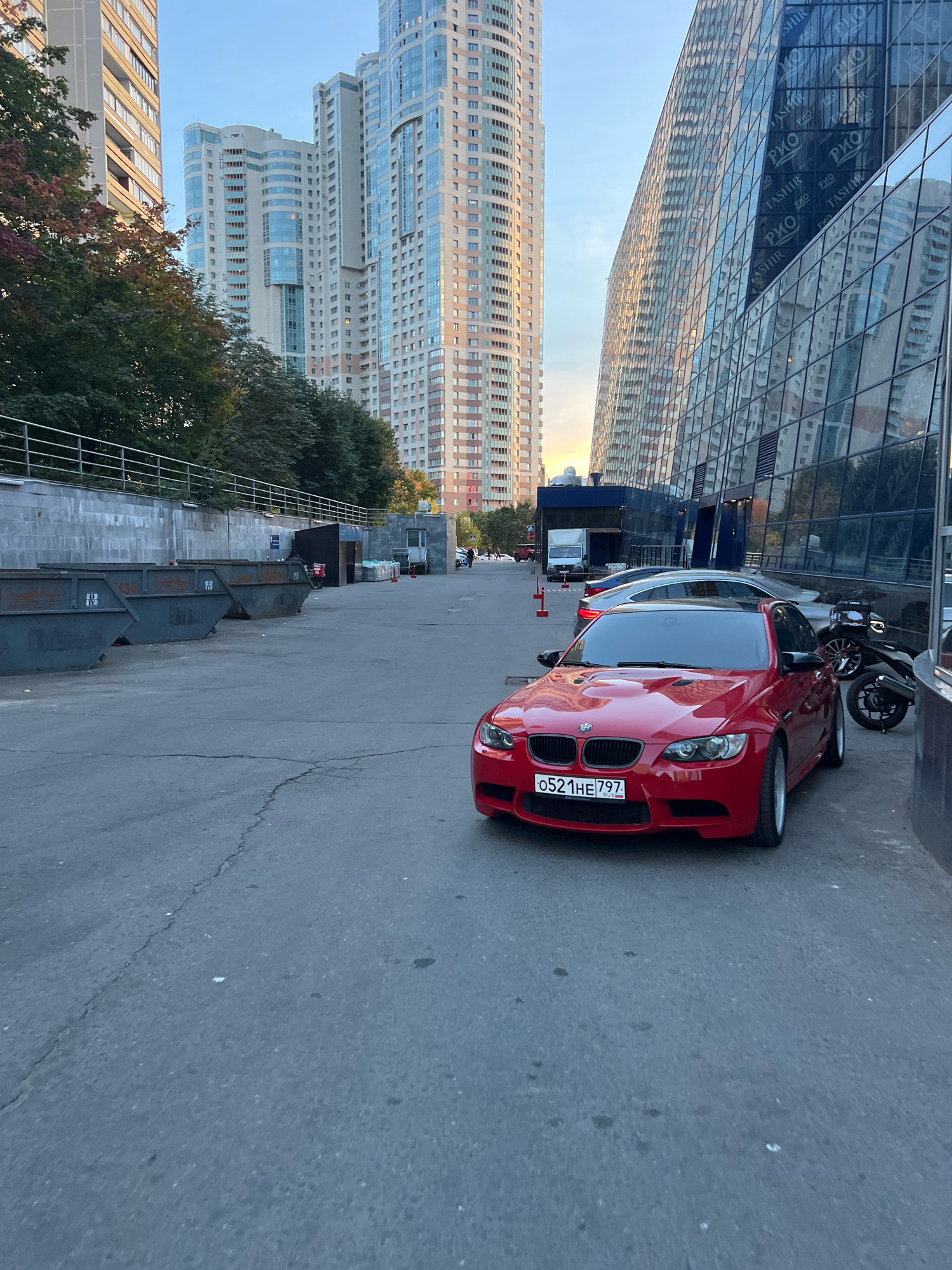 The image depicts an urban scene in Moscow, Russia, likely in a commercial or residential district. The foreground is dominated by a red BMW M3 parked on asphalt. The car has a Russian license plate that reads "0521HE 797". Behind the BMW, a silver car and a black motorcycle are partially visible.
In the background, several tall, modern buildings rise into the sky. One building on the right is characterized by a glass facade that reflects the surroundings. On the left, a row of large industrial dumpsters is lined up against a concrete wall, suggesting a service or utility area. Trees with autumnal foliage are present between the dumpsters and the buildings. The sky is clear and exhibits the warm tones of either early morning or late afternoon. The overall impression is one of a functional urban environment with elements of both modern architecture and practical infrastructure.