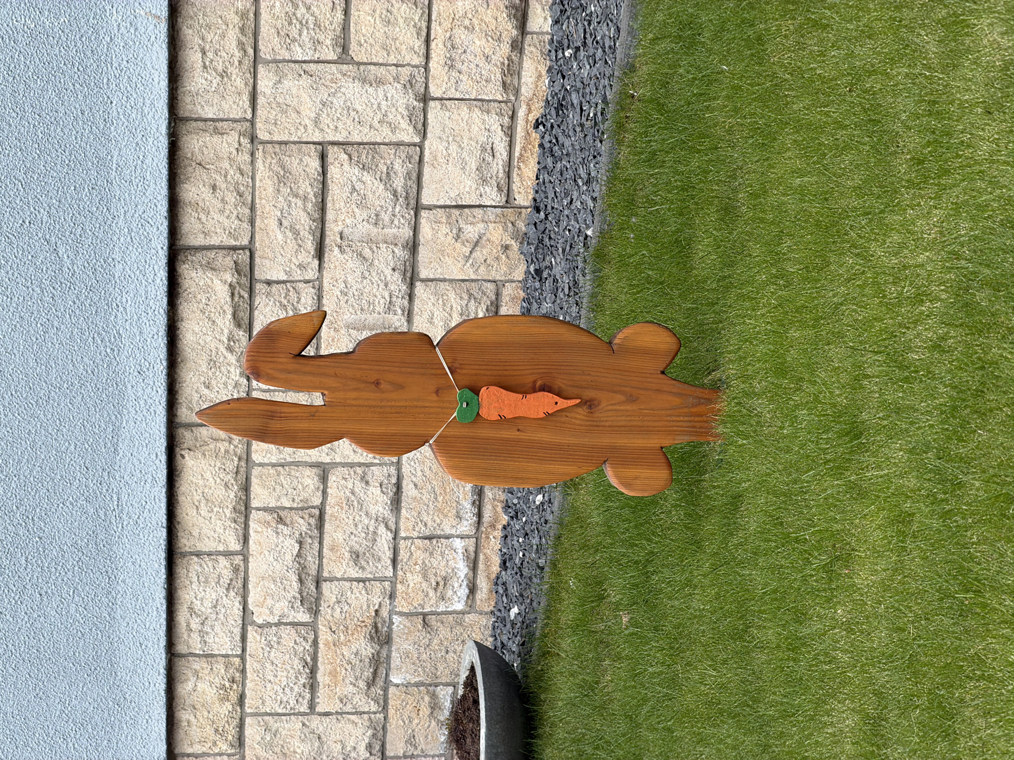 The image is a top-down view of a wooden rabbit decoration with a felt carrot on its back, placed on a stone-paved surface that transitions to a grassy area, bordered by dark gravel. To the left is a light-blue wall. The location is Esch-sur-Alzette, Luxembourg.