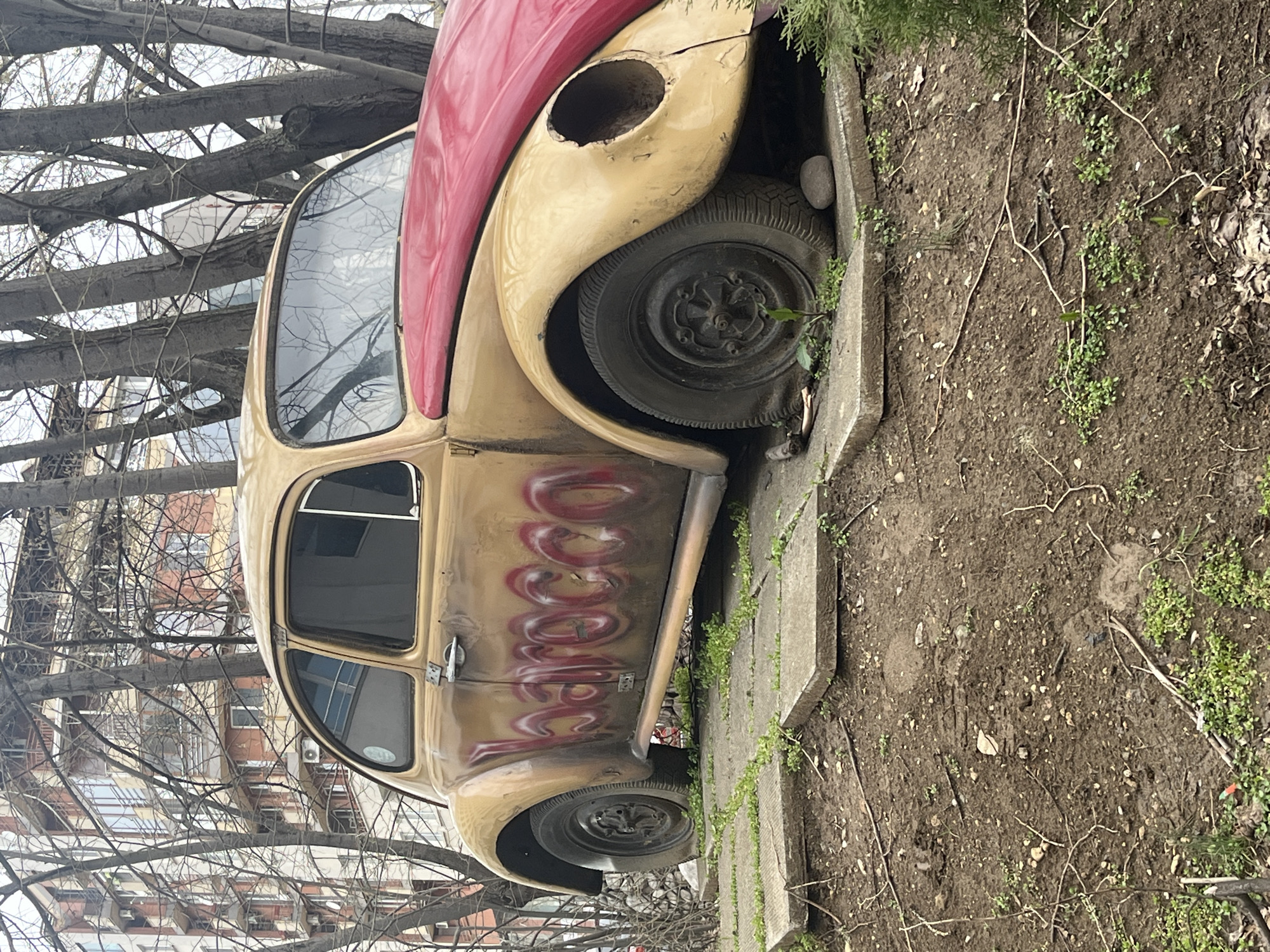 The image shows a partially deteriorated, vintage Volkswagen Beetle parked adjacent to a curb. The car has a two-tone paint scheme with a red roof and a beige body. The front headlight assembly is missing. Graffiti is visible on the side of the car. The vehicle is positioned on a concrete slab or walkway, which is adjacent to a dirt and grassy area. Bare tree branches are visible in the background, and buildings suggest an urban setting. Based on visual cues and commonly found landmarks, this is very likely within the city of Sofia, Bulgaria.