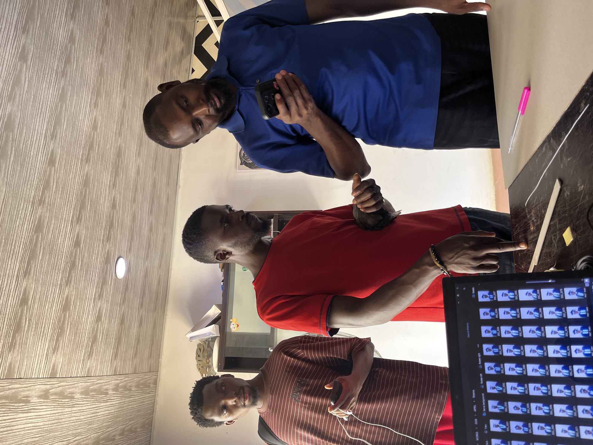 Three adult males are in a room with patterned wallpaper. The man at the top of the frame wears a blue shirt and holds a black mobile phone. The man in the middle wears a red shirt and points to a computer screen displaying thumbnails of images. The man at the bottom wears a horizontally striped brown shirt. The location is Jos, Nigeria. A pink pen lies on a dark table. A white cable runs from the table to the striped shirt. There is a cabinet with items inside behind the red shirt. The ceiling contains a light fixture.