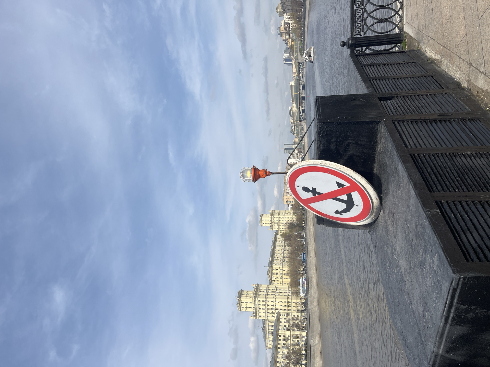 An eye-level, outdoor, full shot depicts a “No Anchoring” sign on the Moscow River embankment, Moscow, Russia, under a blue, cloudy sky.
The sign is circular with a white background and a red border, depicting a black anchor with a red diagonal line through it. The sign is affixed to a black, waist-high structure on the river embankment. To the left of the sign is a pole with an orange lamp enclosed in a metal wire cage.
In the background, across the river, are several large, light-colored buildings lining the embankment. Trees are sparse, some showing bare branches. Several small boats are visible on the water.
The embankment is grey, wet, and reflects the sky and buildings. A black, ornamental fence runs along the edge of the embankment.