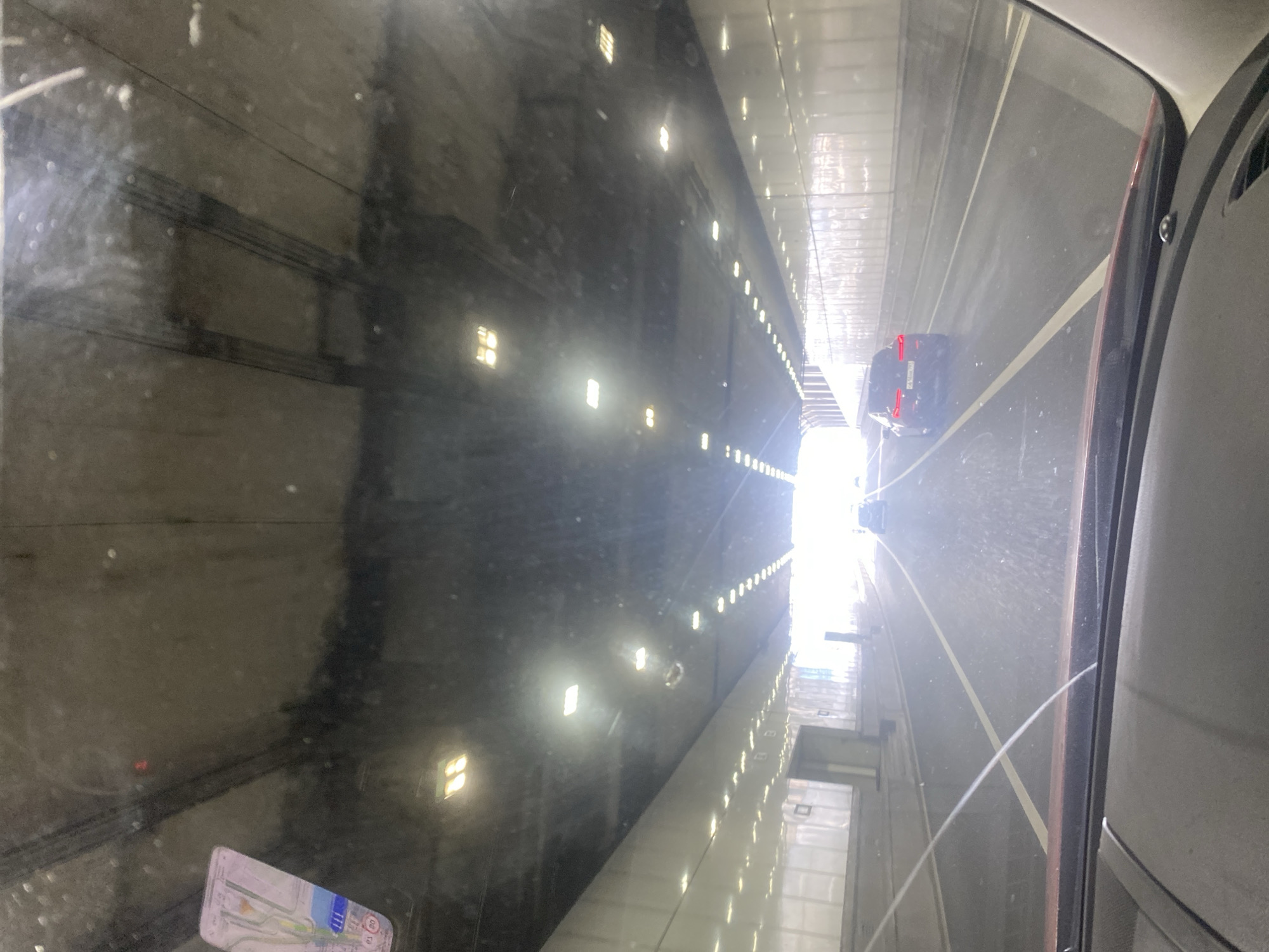 The image shows the interior of a car driving through a tunnel in Moscow, Russia. The driver's perspective is from behind the windshield, which is dirty with streaks and dust. In the foreground, the car's dashboard and windshield wipers are visible. The tunnel is illuminated by a series of lights on the ceiling. The tunnel walls are white and reflective. There are other cars in the tunnel, including a dark-colored sedan in front of the car. The road is wet, and there are lane markings on the asphalt. The exit of the tunnel is visible in the distance, with bright daylight shining through. A sticker is affixed to the upper left corner of the windshield.